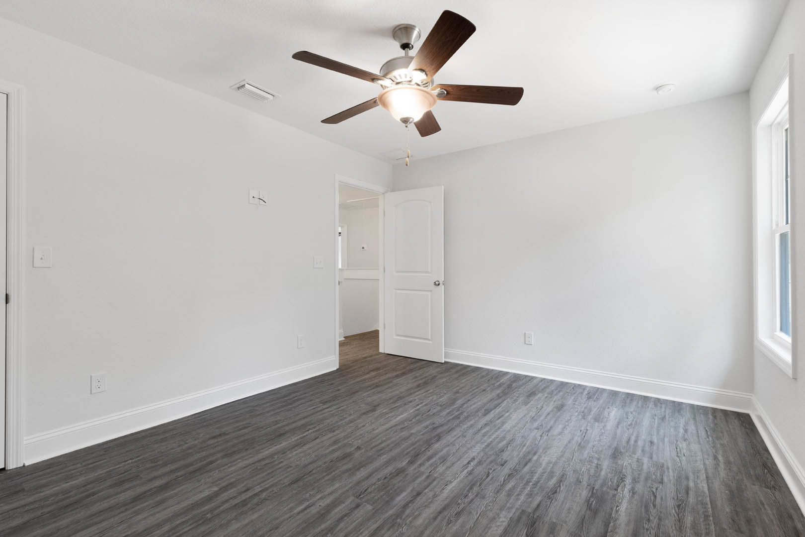 Ceiling fan with light fixture mounted on white ceiling above grey wood flooring, white door with silver knob, neutral walls.