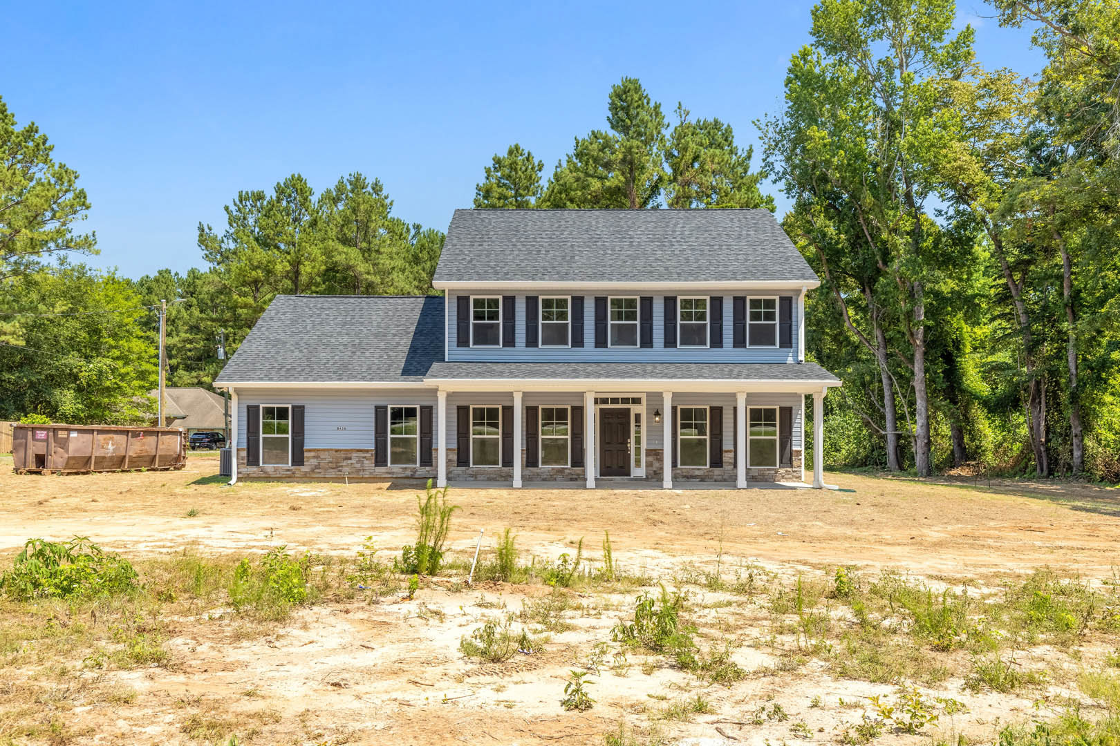 Two-story house with beige siding, covered front porch, black door with white frame, white-trimmed windows, dirt yard in foreground, mature trees in background