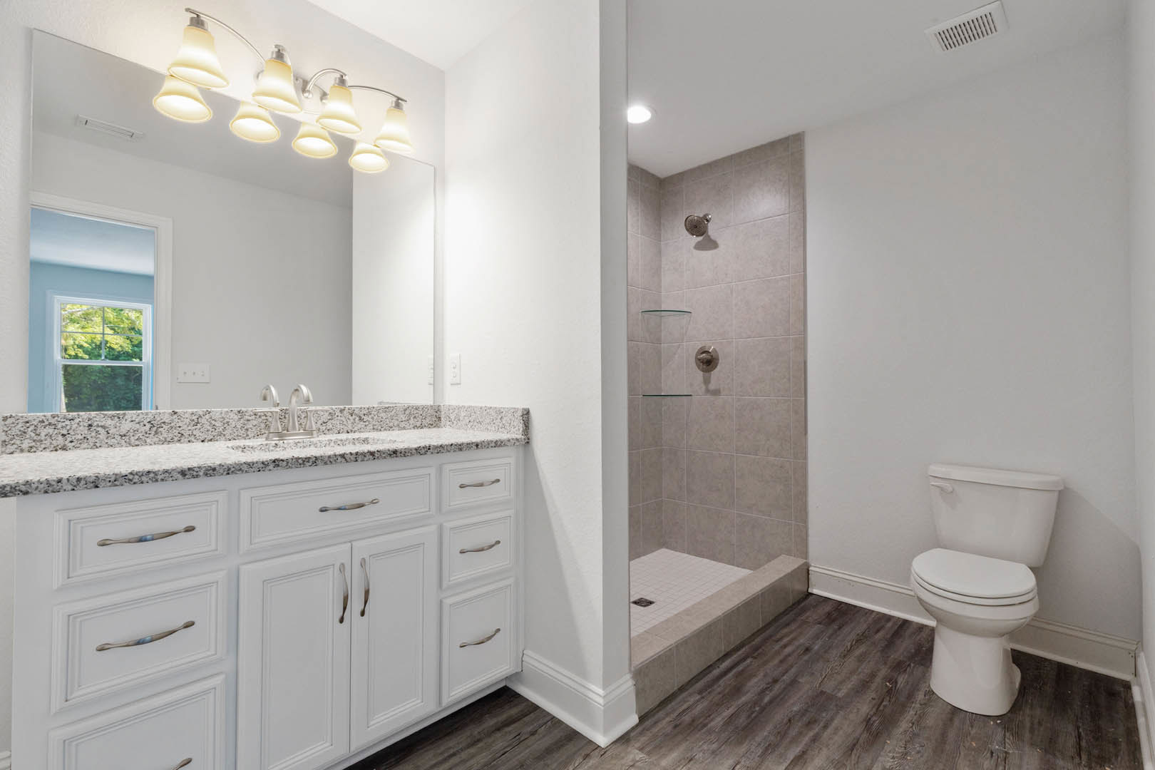 Modern bathroom featuring a glass-enclosed shower, white toilet with closed lid, marble countertop vanity, overhead row of lights, and window overlooking trees