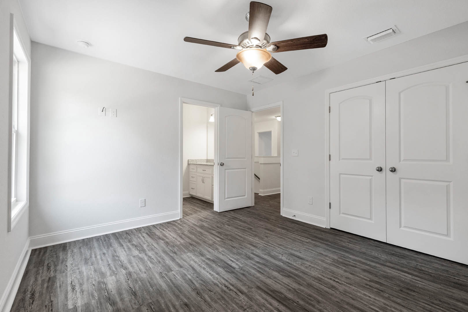 White-walled room with wood flooring, ceiling fan with light fixture, white double doors, and a white door featuring a silver knob and window panel