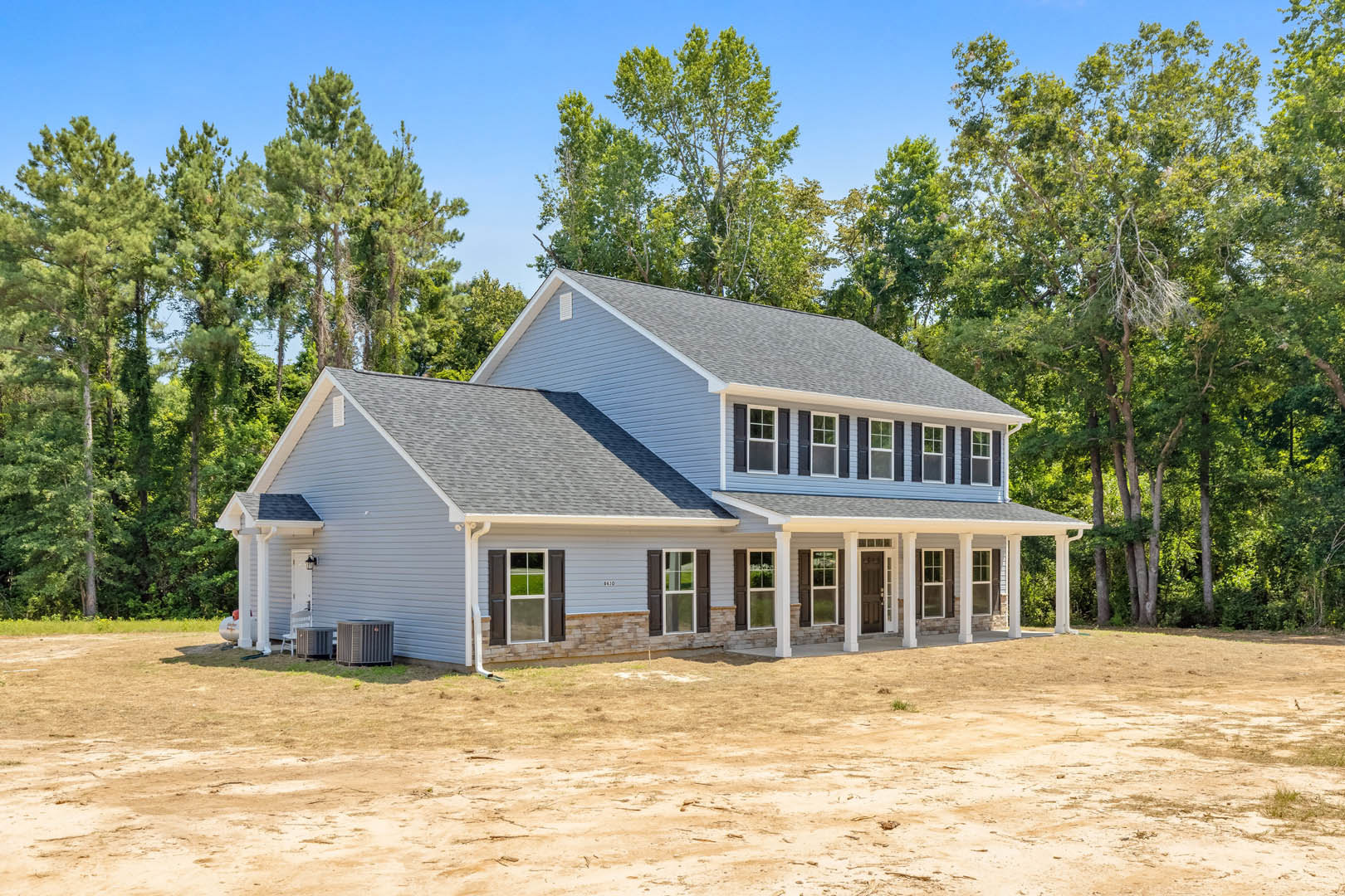 Blue metal roof house surrounded by mature trees, dirt yard in foreground, white-framed windows, gray siding, red sign near entrance
