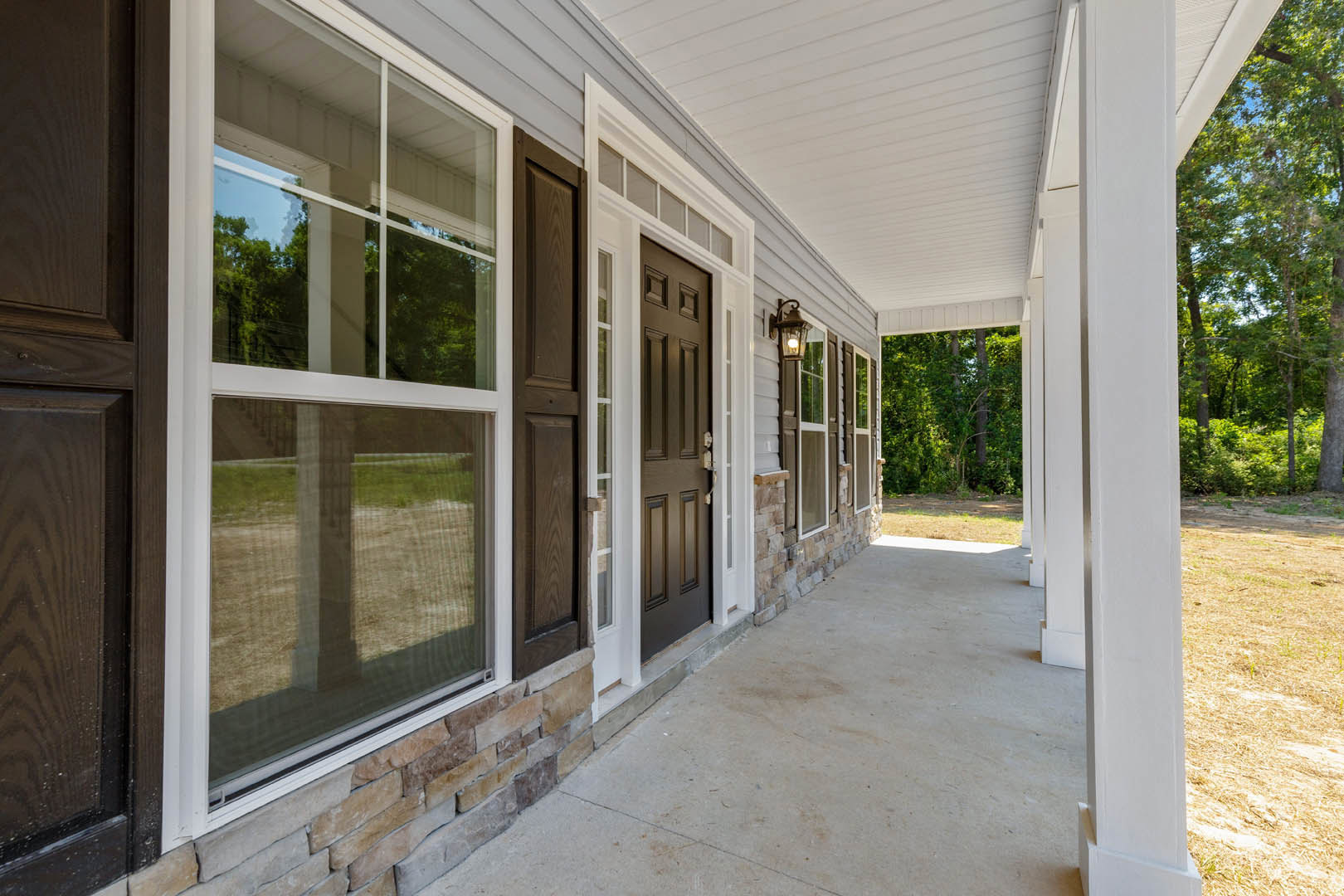 Front porch with stone walls, white-framed windows, shaded by trees, composite flooring, and wooden door