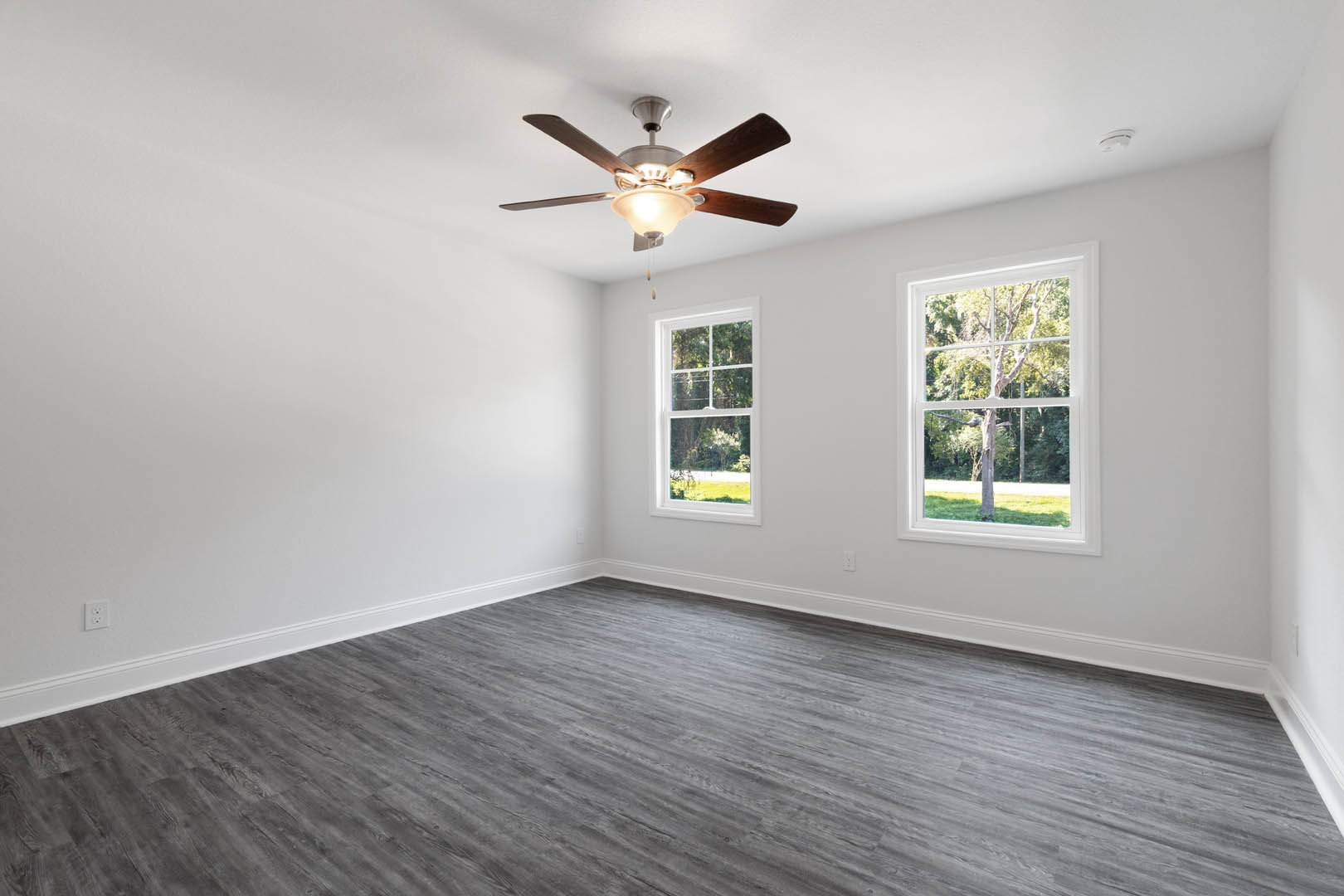 Living room with wood flooring, plaster walls, ceiling fan with light fixture, large windows overlooking trees and forest.