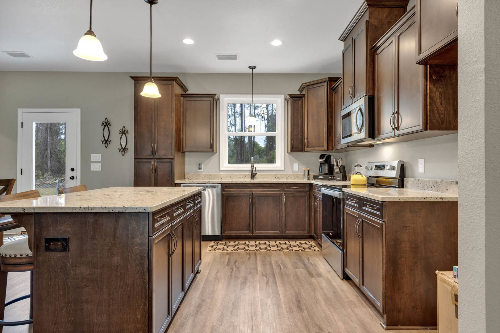 Kitchen with dark wood cabinets, wood flooring, marble-topped island, yellow tea kettle on the counter, built-in microwave, white door with window, and large window letting in