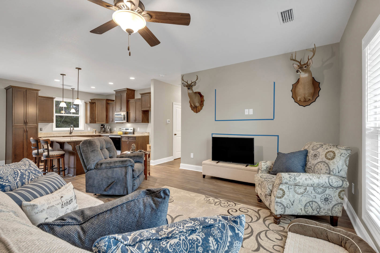 Living room with grey recliner, mounted deer head on wall, couch with pillow, flat-screen TV displaying blue stripe, ceiling fan with light fixture