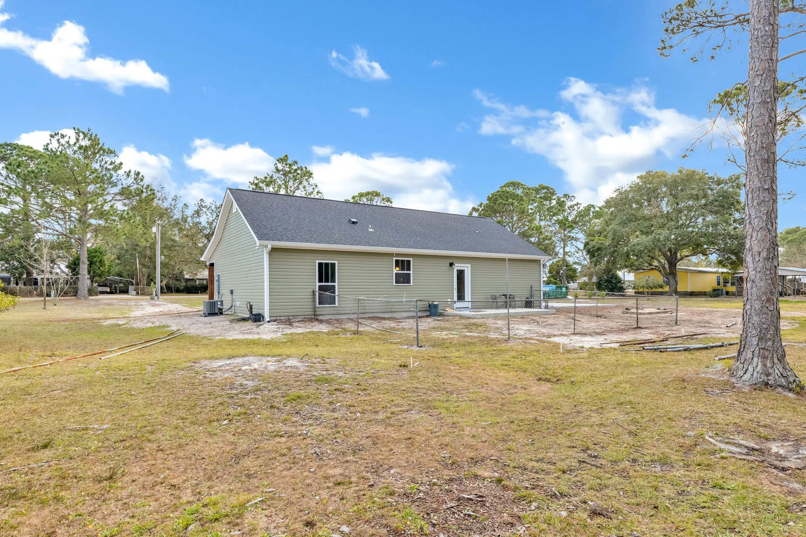 White farmhouse with gabled roof, fenced grassy yard, mature trees, and large front windows under a partly cloudy sky
