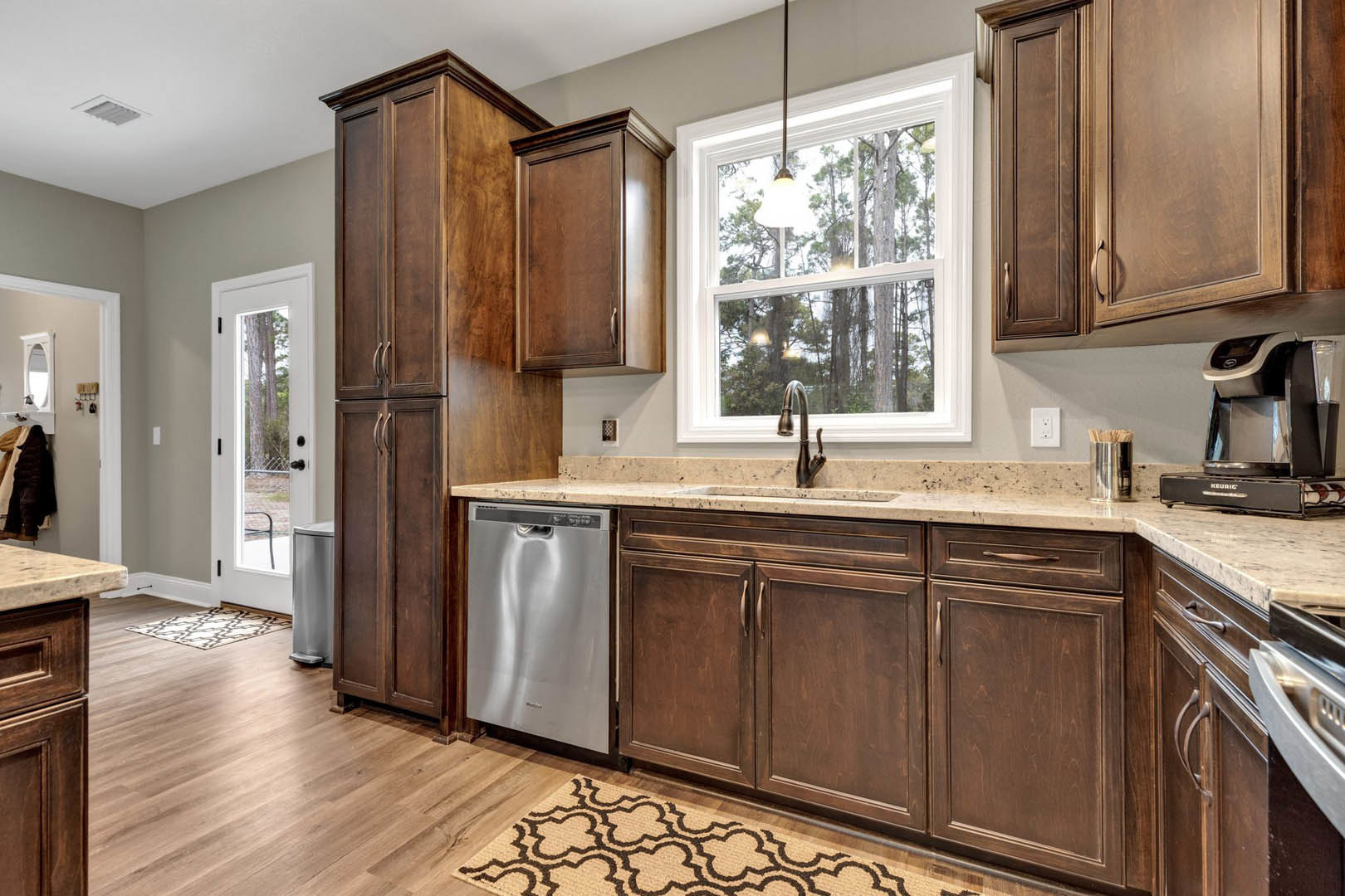 Dark wood kitchen cabinets, stainless steel dishwasher, white fabric chair with brown frame, window above sink and faucet, close-up views of cabinetry and rug