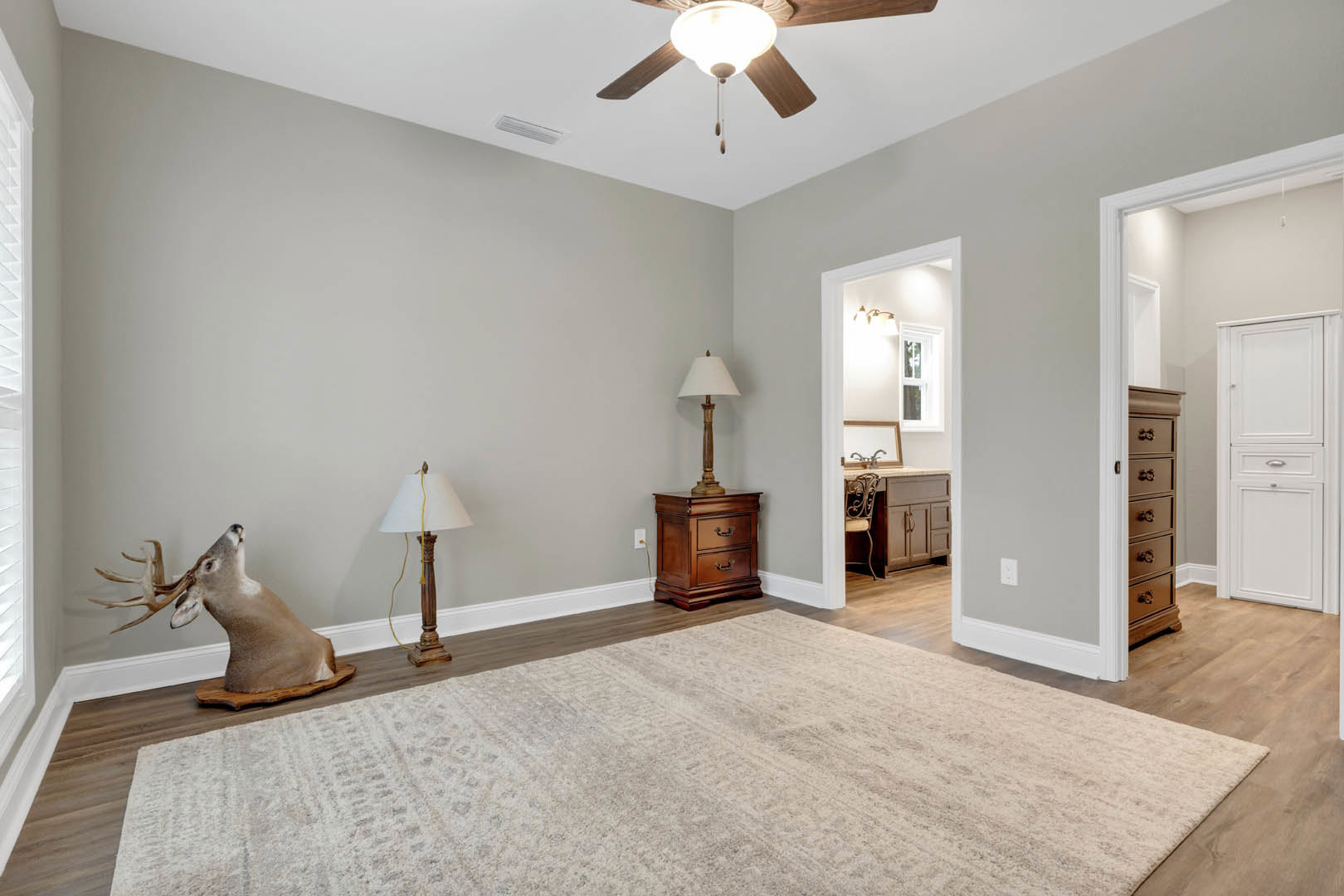 Living room with laminate flooring, ceiling fan, white cabinet with drawers, wooden nightstand topped with a vase, deer head with antlers mounted on the wall, lamp with white