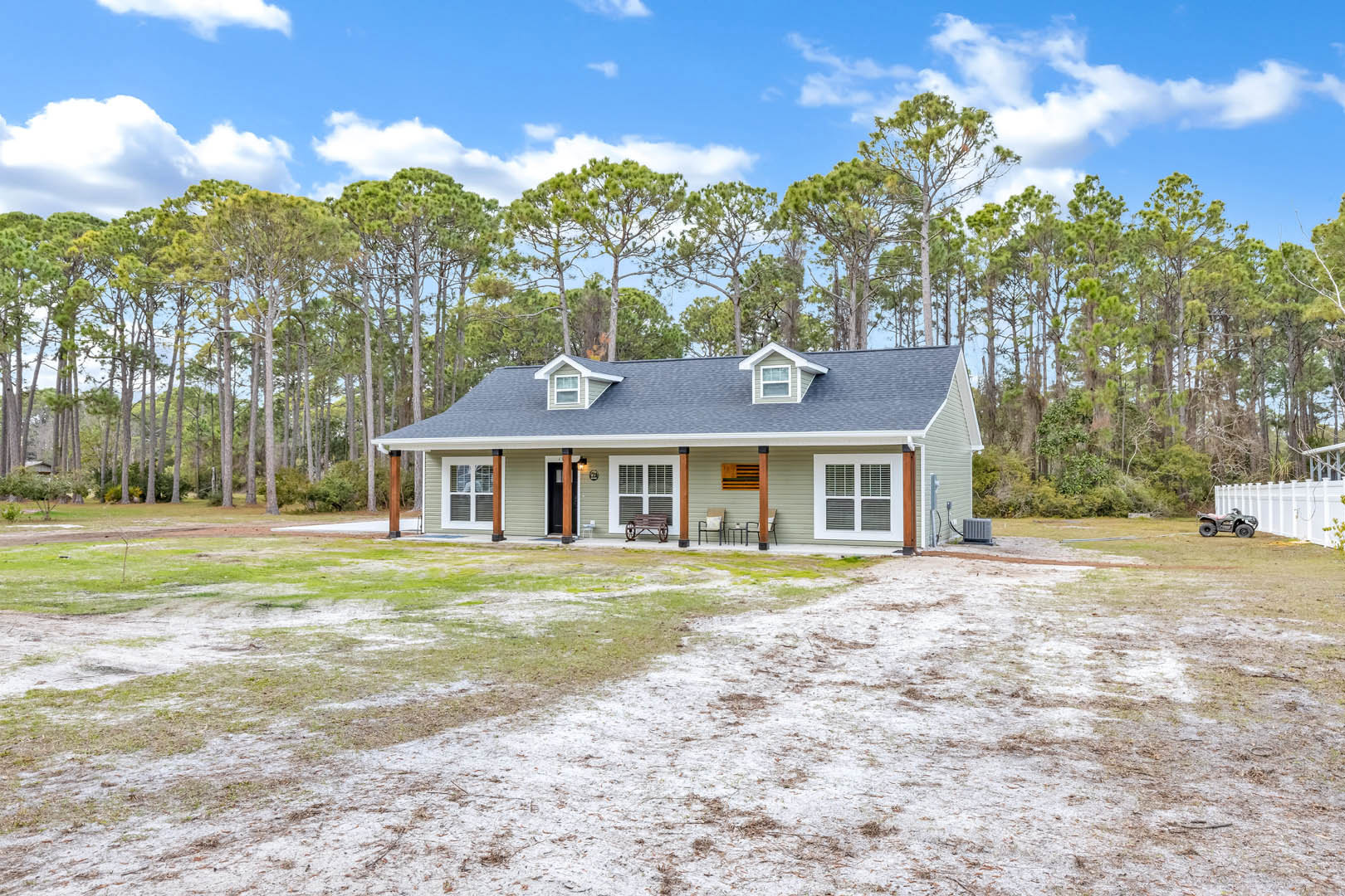 Two-story home with white siding, large windows, front porch featuring a bench and chairs, manicured lawn, mature trees in the background, and blue sky with scattered clouds