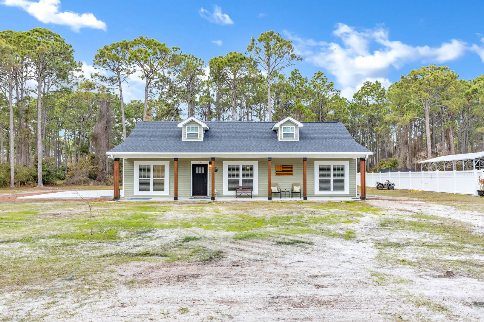 White house with black front door, white trim, covered porch, wooden bench with metal frame, dirt yard, mature trees in background, cloudy sky