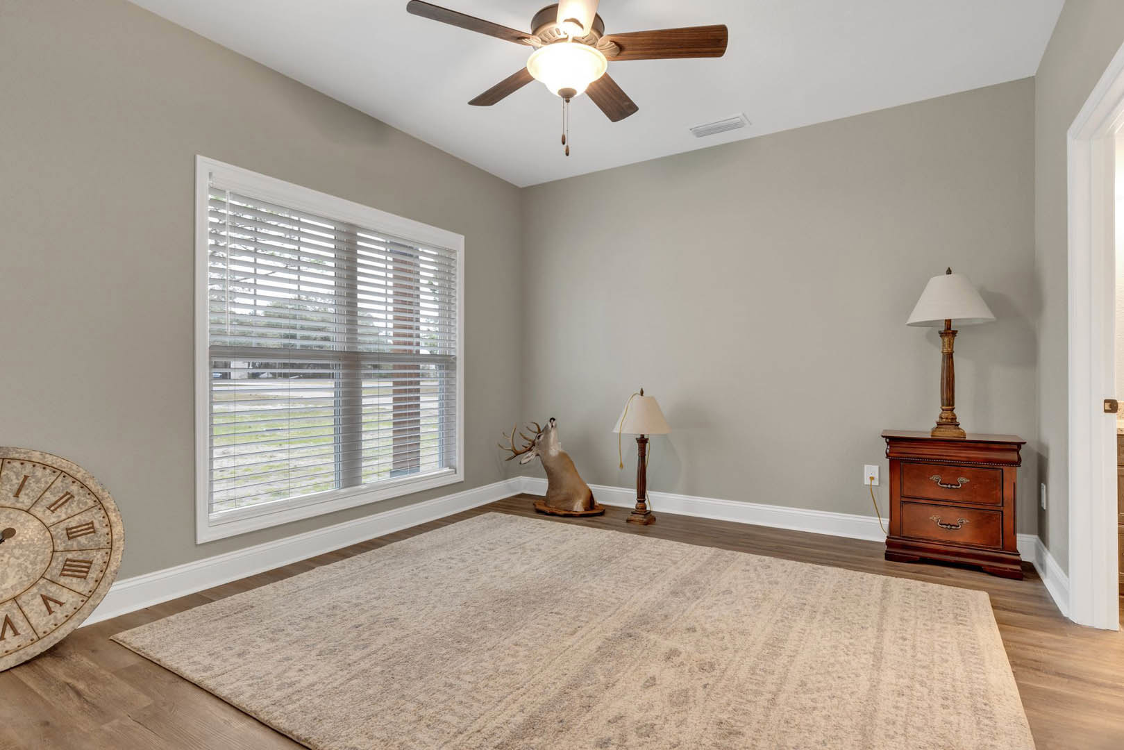 Living room with hardwood floors, ceiling fan with light fixture, mounted deer head above a rug, dresser with vase, window with white blinds