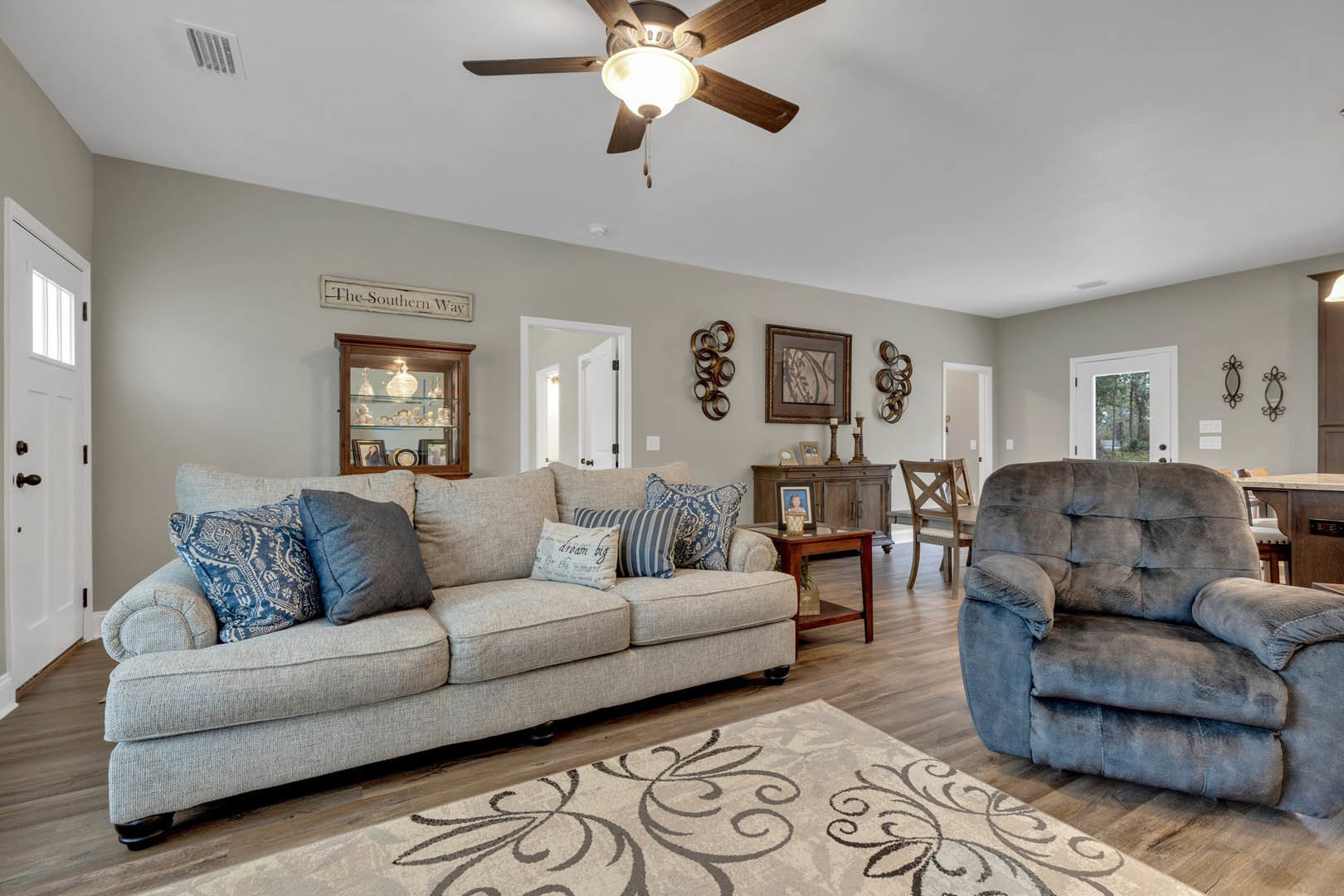 Living room with a gray couch topped with pillows, ceiling fan with light fixture, glass cabinet displaying decorative objects, framed artwork on wall, and a gray accent chair.