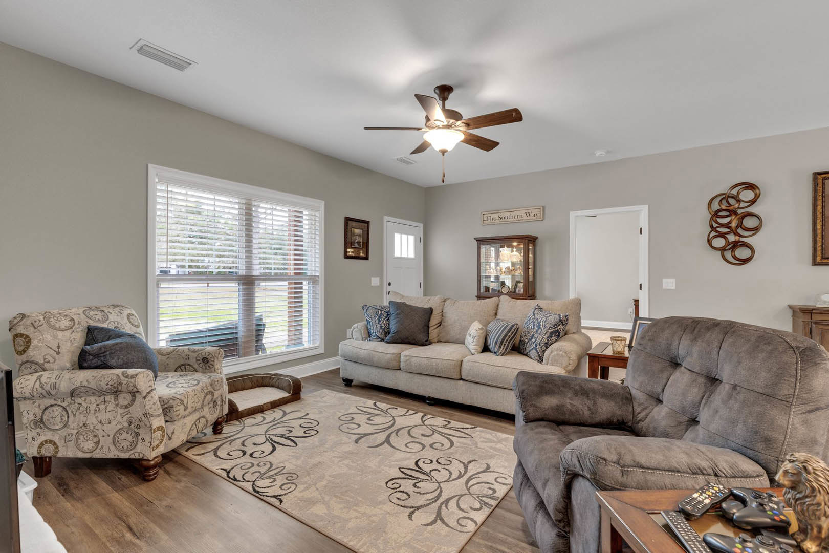 Living room with beige couches, accent pillows, wooden coffee table holding remote controls, upholstered chair, ceiling fan with light fixture, window with white blinds, neutral