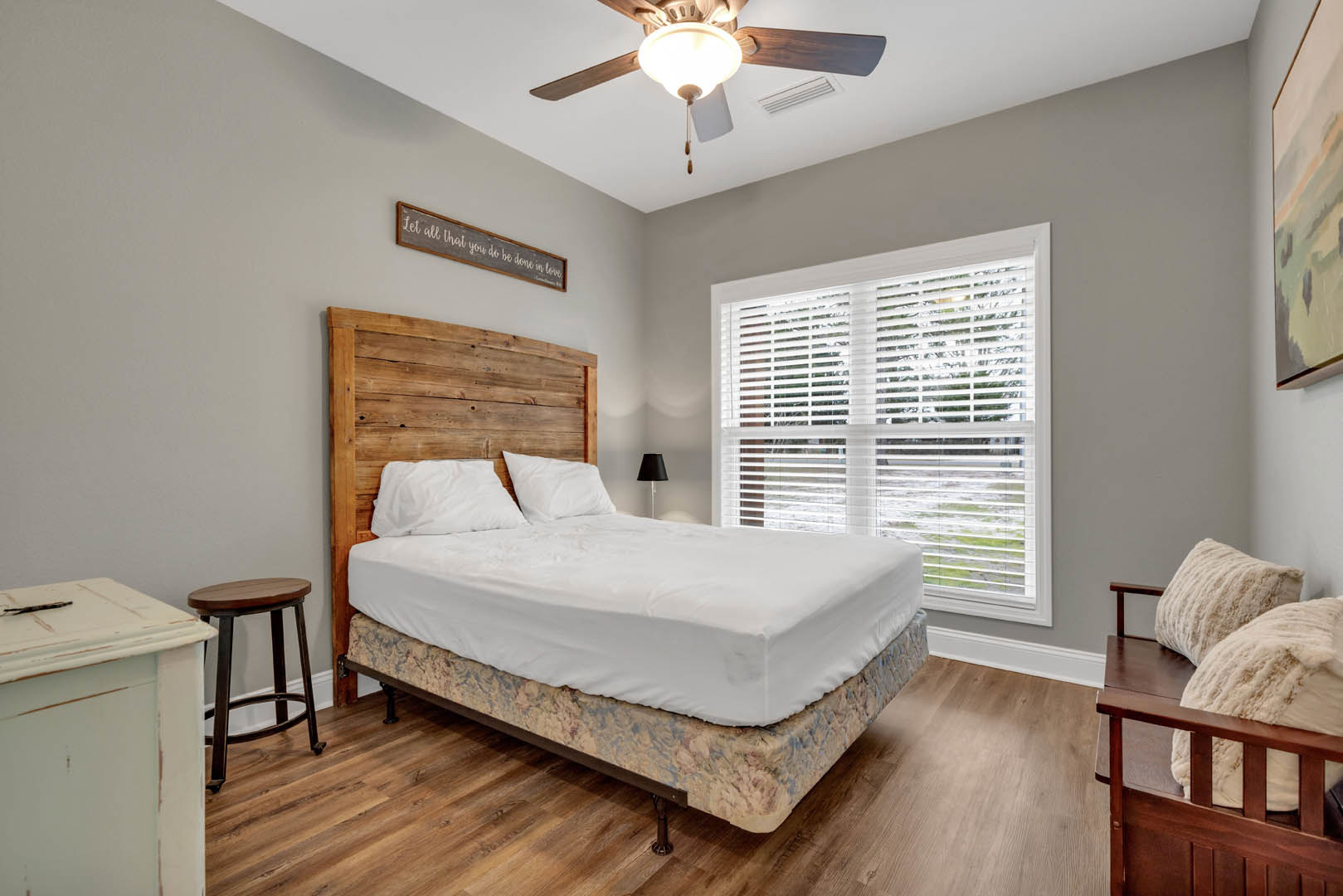 Bedroom with wood flooring, ceiling fan, bed featuring a wood headboard, nightstand with lamp, window with white blinds, wall-mounted sign, upholstered stool, and framed painting.