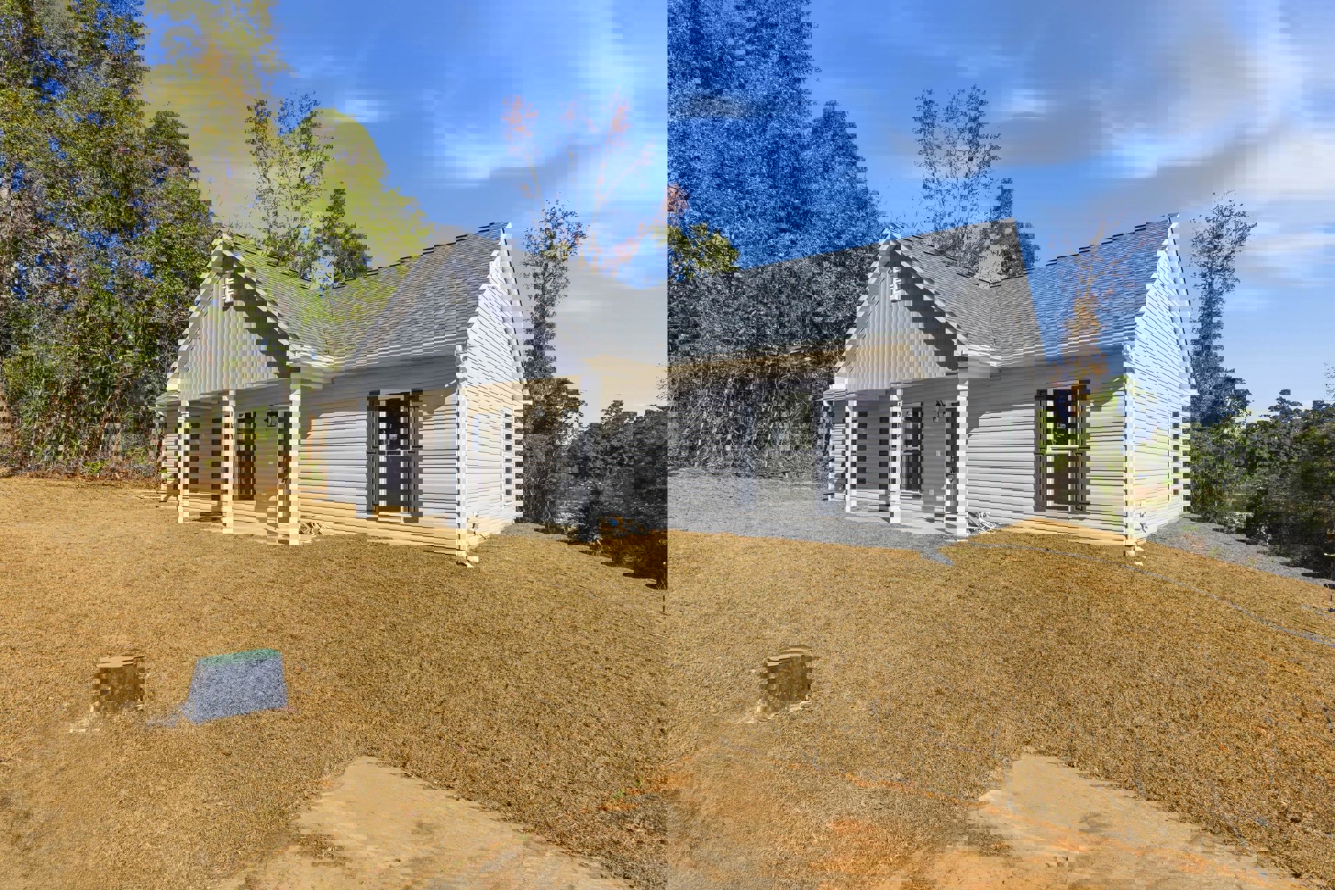 Two-story house with white siding, covered porch, and large windows, set on a green lawn with a concrete sidewalk; mature trees and Robert Frost Farm visible in the background.