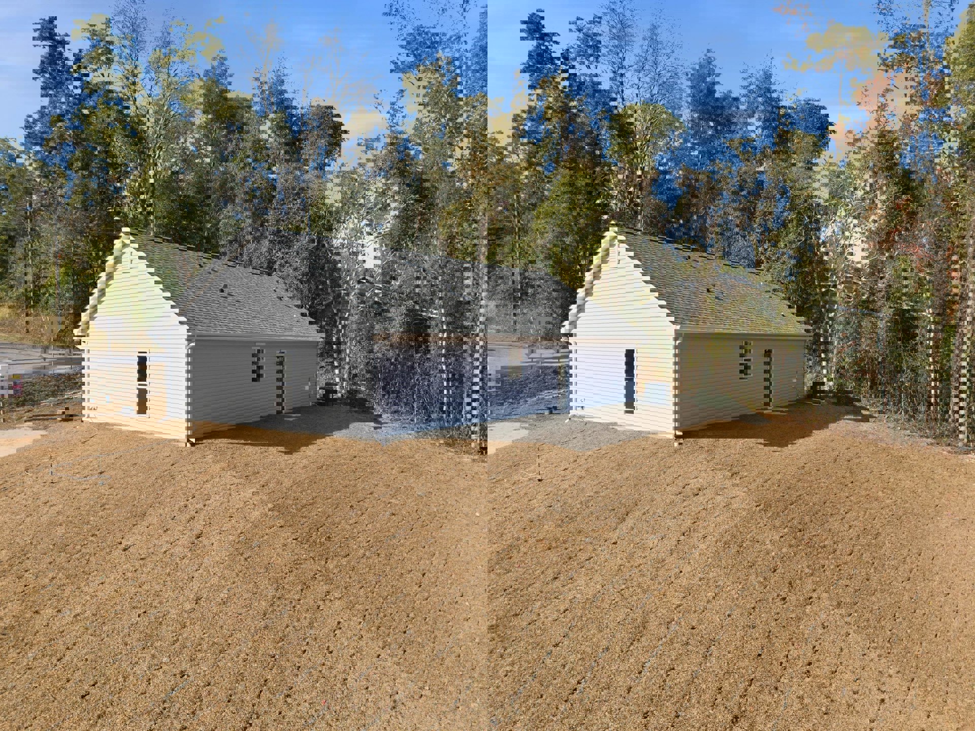 White house with attached garage and window, surrounded by a large patch of bare dirt; trees in the background, sign on a pole near the property, roof features two vents.