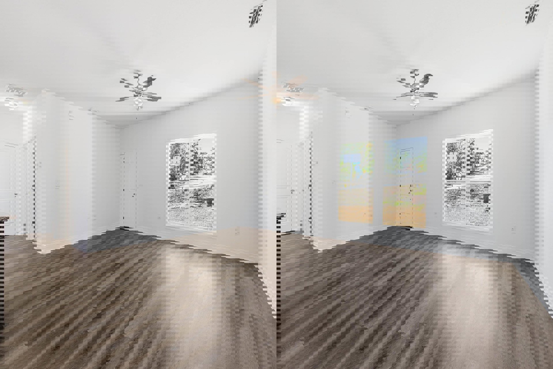 Wood flooring, white walls, ceiling fan with light fixture, white door with silver handle, large window overlooking street and trees