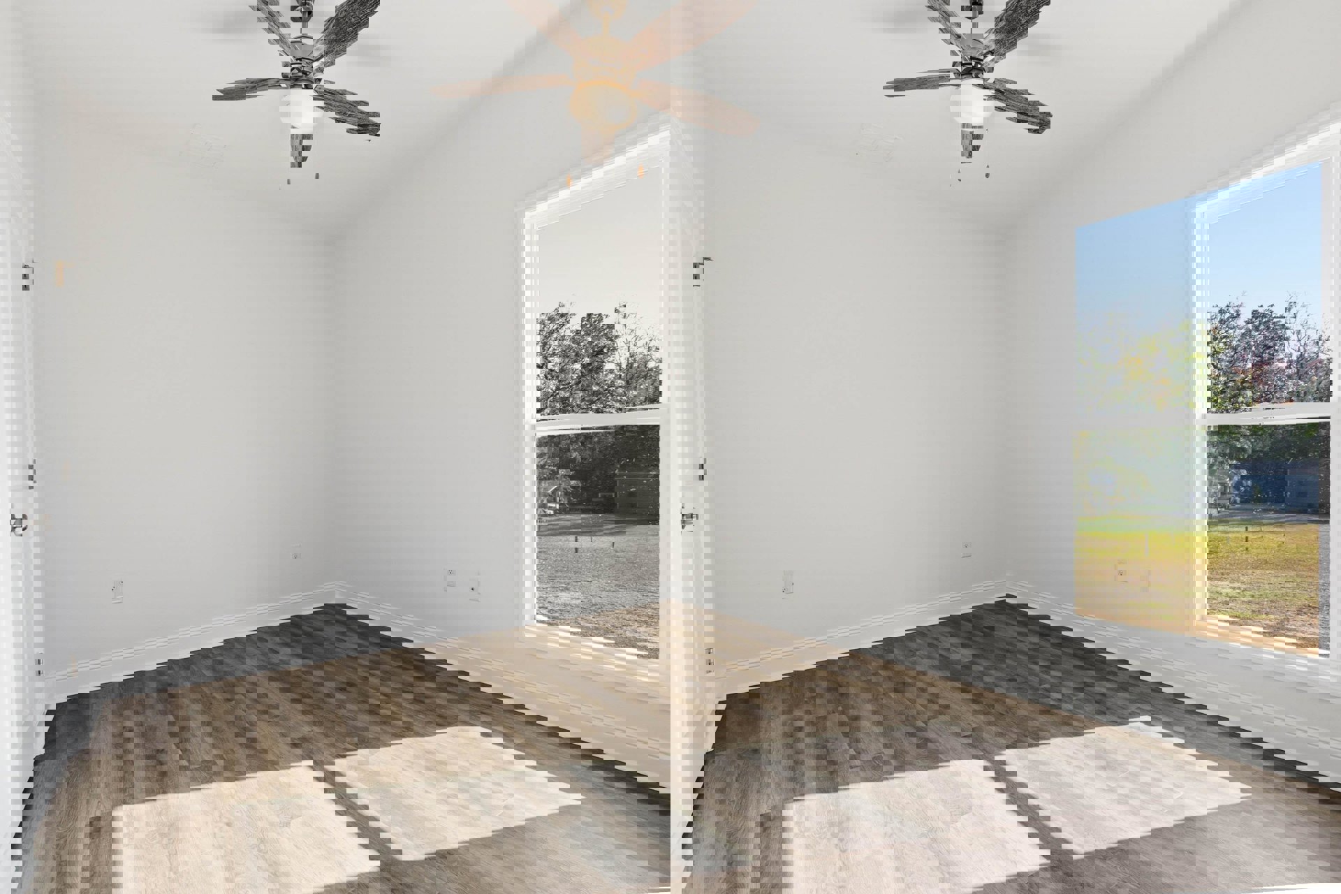 Wood flooring and white walls in a bright room with a ceiling fan, large window overlooking a grassy fenced yard and trees