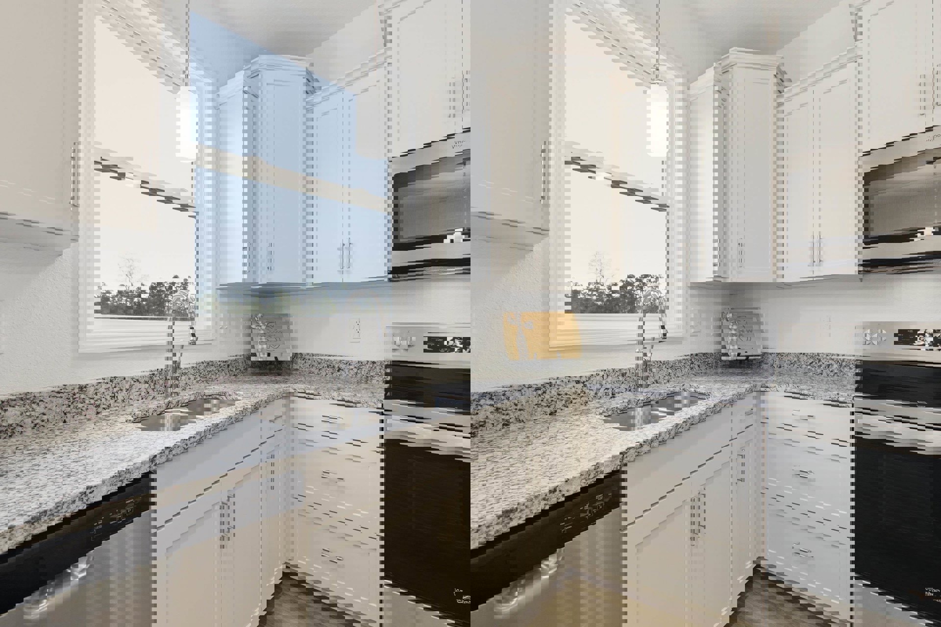 White kitchen cabinets with granite countertops, stainless steel oven and microwave, undermount sink, sunlight streaming through window, wooden cutting board with bow on counter.