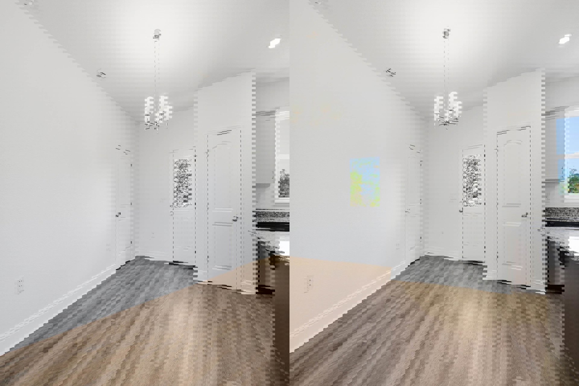 White walls, wood flooring, white doors with silver knobs, ceiling light fixture, window showing trees outside, partial view of kitchen cabinetry and countertops