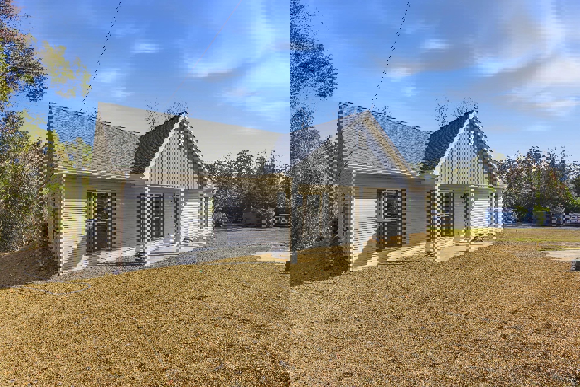 Two-story house with white siding, covered front porch, large windows, manicured lawn, mature trees in the background, and blue sky with scattered clouds