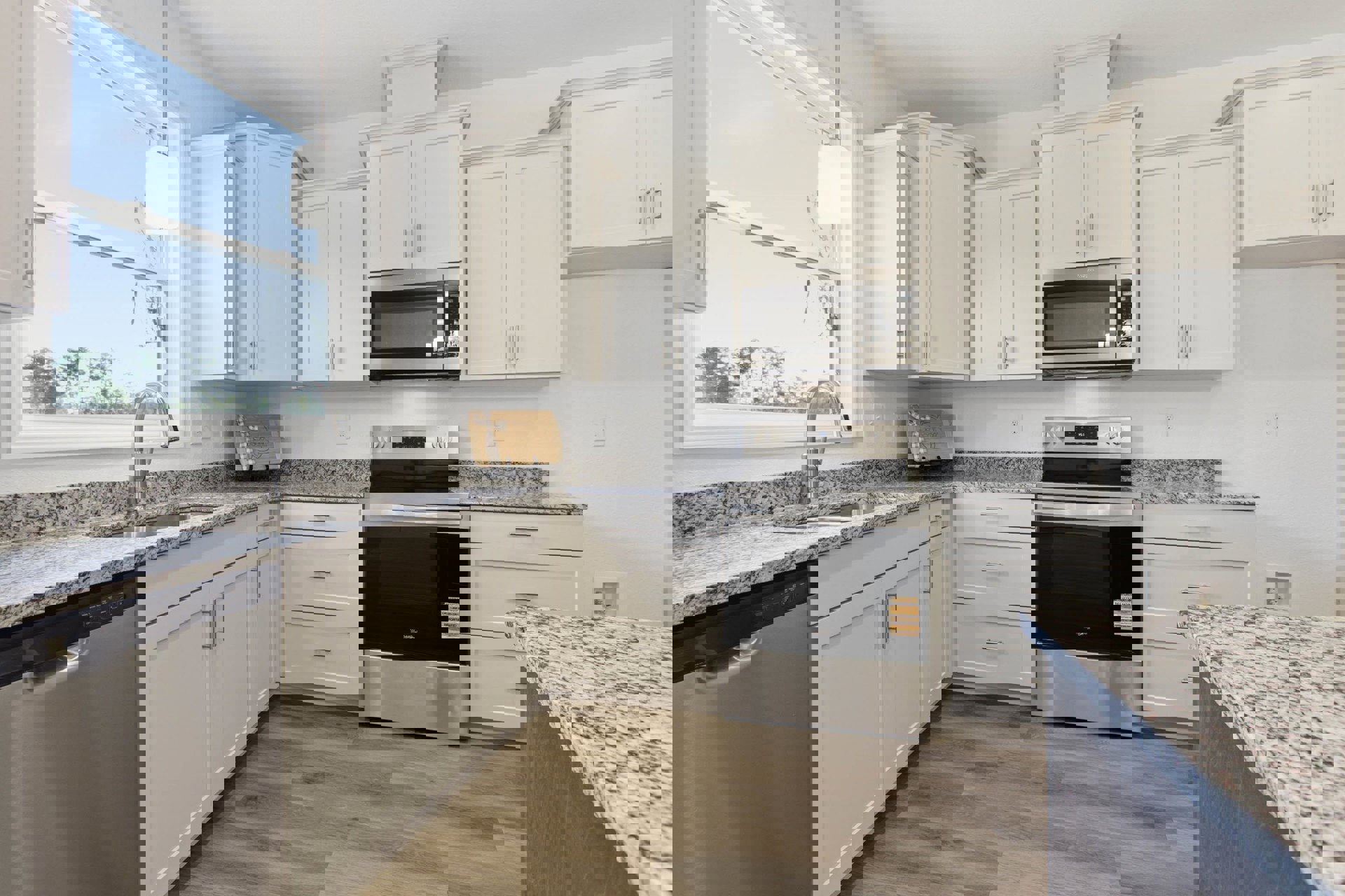 White kitchen cabinets and speckled granite countertops, stainless steel oven with black glass door, open microwave, white window with overhead light fixture, wooden box with bow