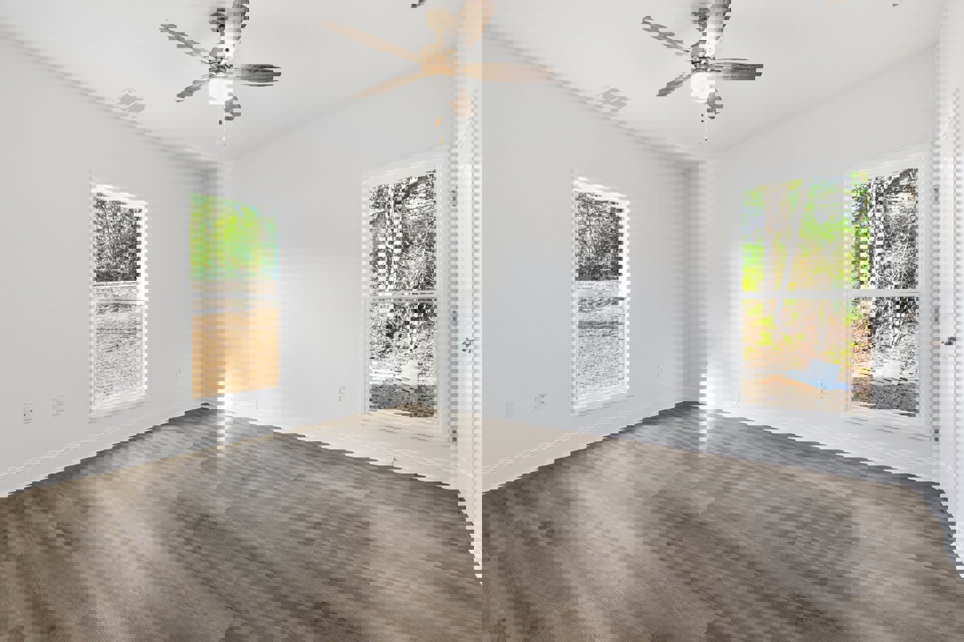 Ceiling fan with light fixture centered above wood flooring, two large windows with views of trees and yard, white plaster walls and ceiling