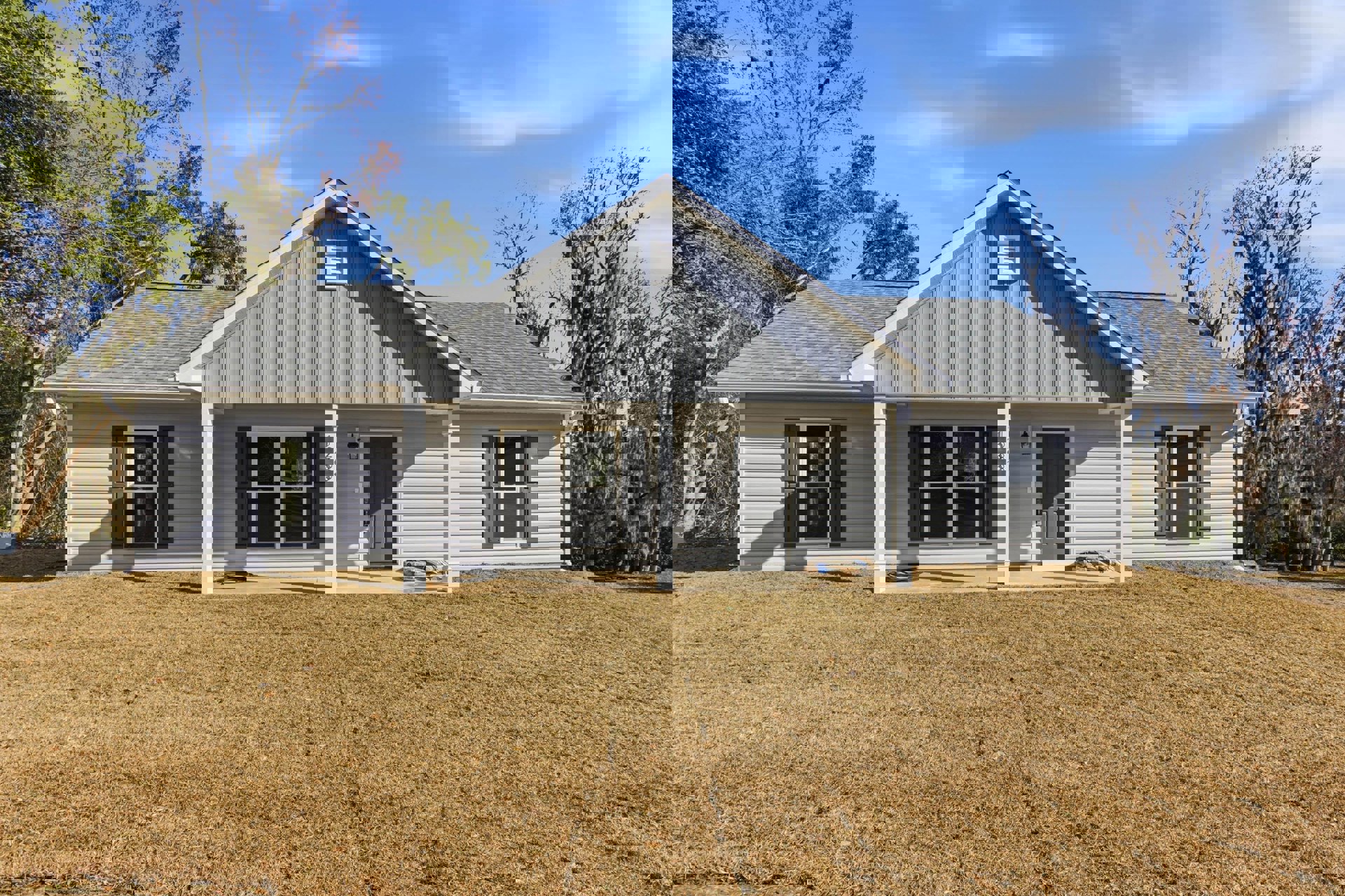 White-walled house with grey front door, large windows, and gabled roof, surrounded by green lawn and mature trees under a partly cloudy sky