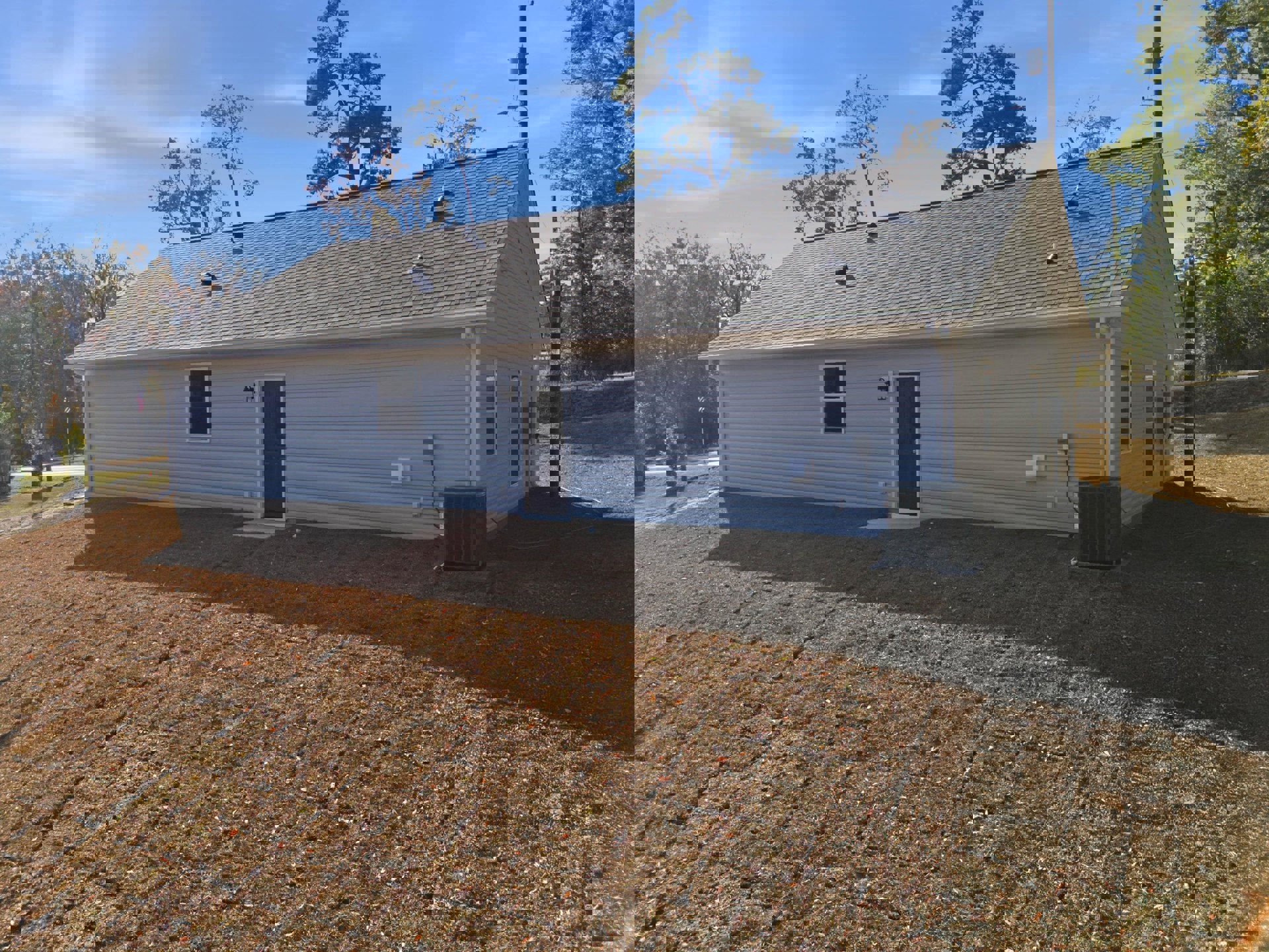 Modern cottage-style home surrounded by mature trees and exposed dirt lot, white-framed windows, covered porch with single entry door, black heat pump unit positioned near exterior