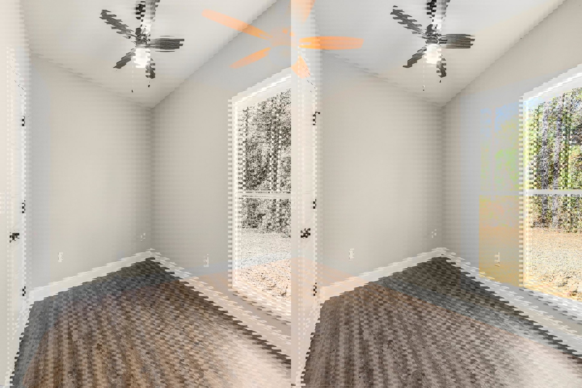 Ceiling fan with light fixture above wood flooring, window overlooking trees, door with black handle, neutral walls