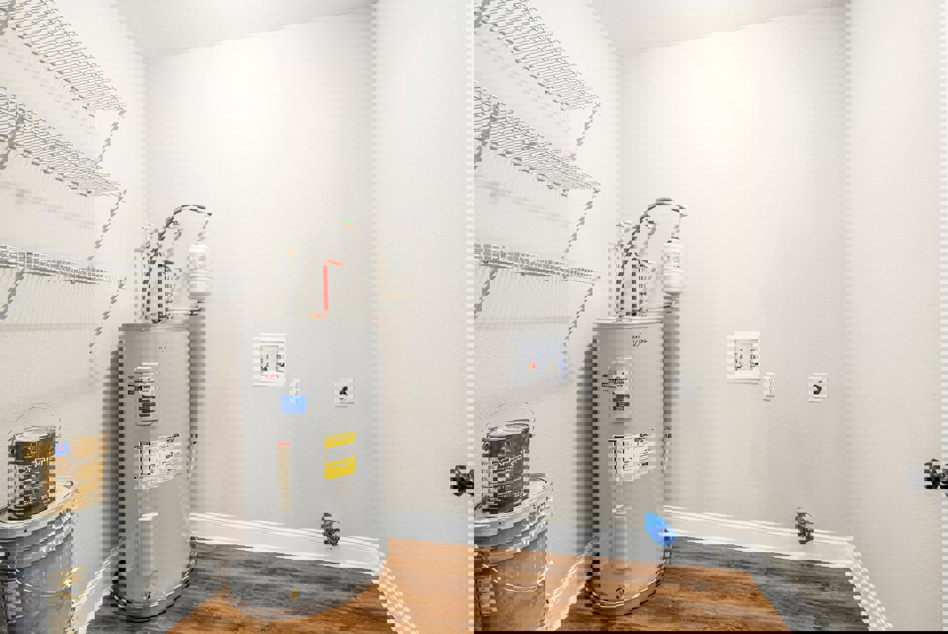 White water heater tank with two valves against a plaster wall, paint cans stacked on a shelf nearby, indoor utility room with visible floor and sink.