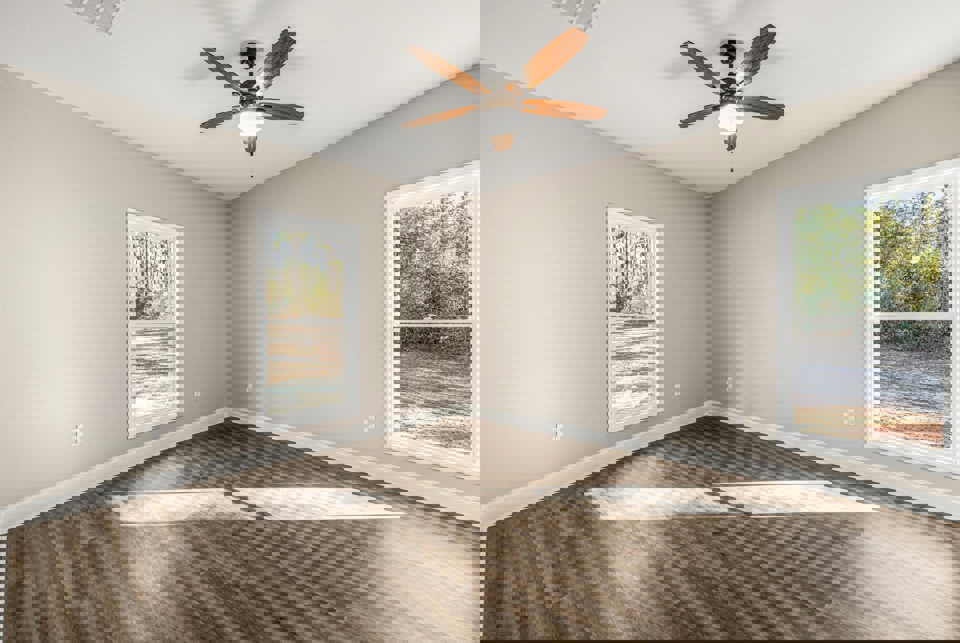 Ceiling fan with light fixture centered above wood flooring, white plaster walls, and large windows revealing leafy trees outside