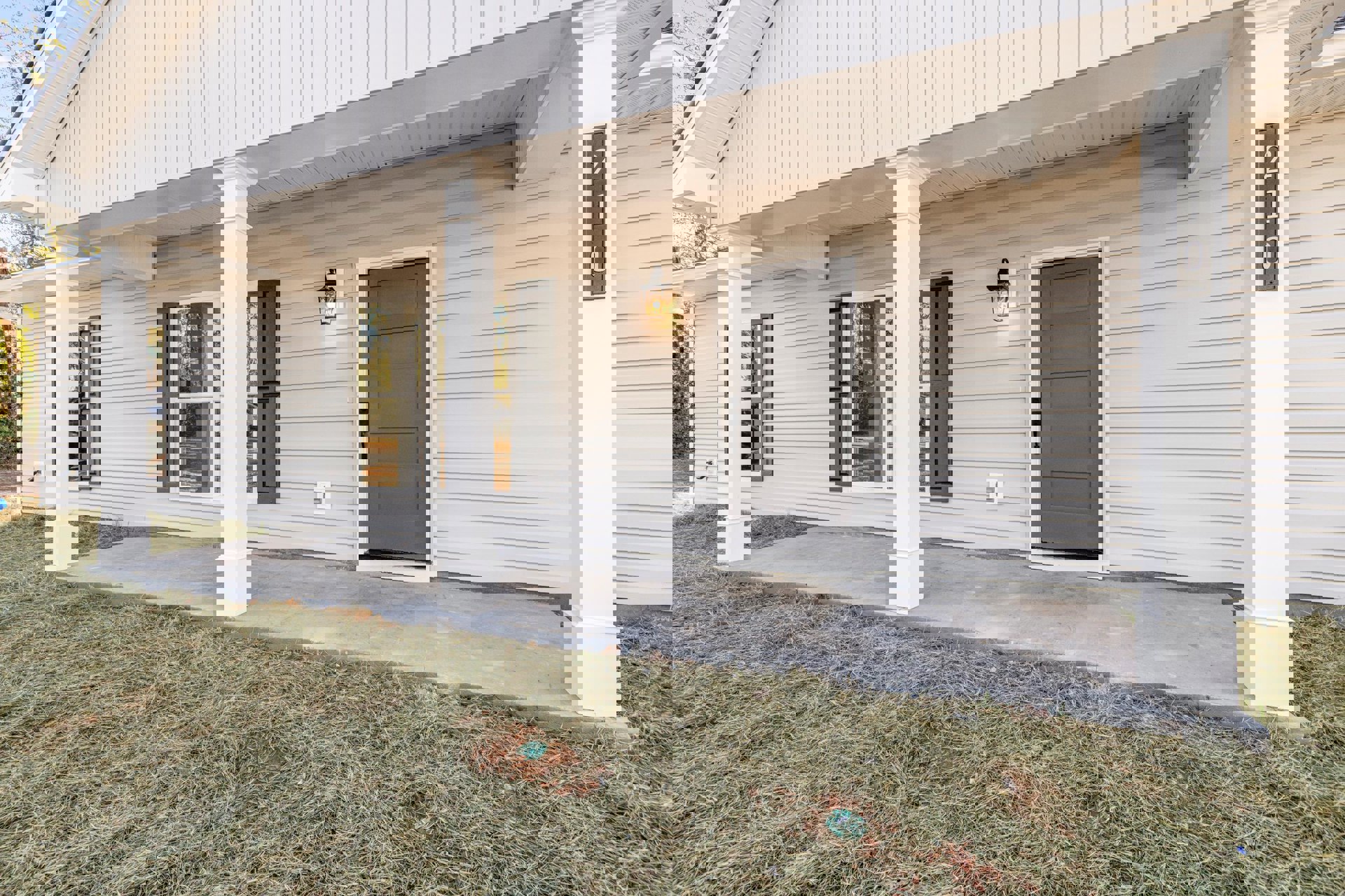 Grey front door with metal handle, white column, rectangular house number sign, wall-mounted light fixture, and green lawn in front of light-colored siding exterior.