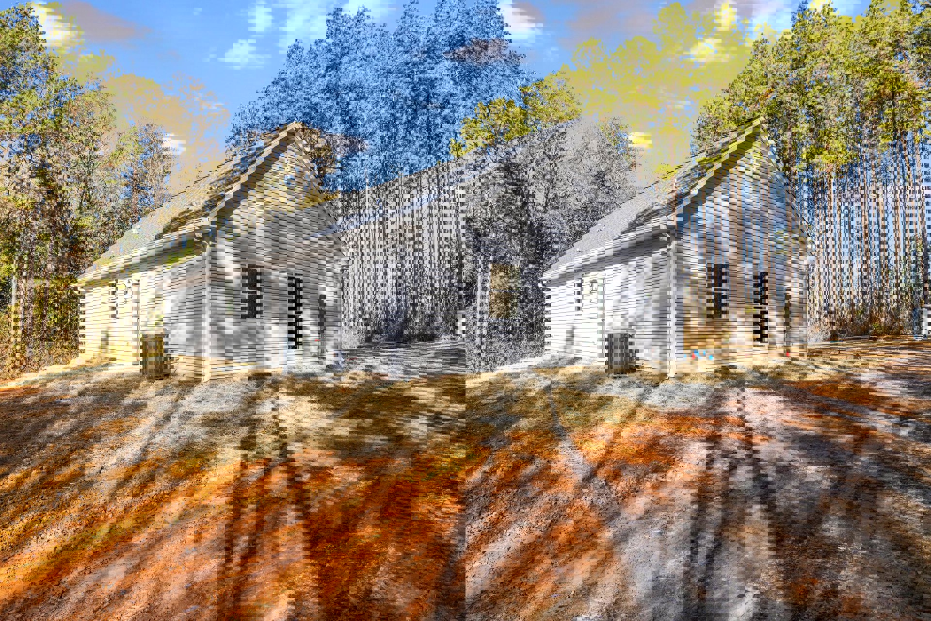 Modern cottage-style home with white-framed windows, surrounded by tall trees and blue sky, tree shadows cast across dirt ground in the foreground, large metal object with red