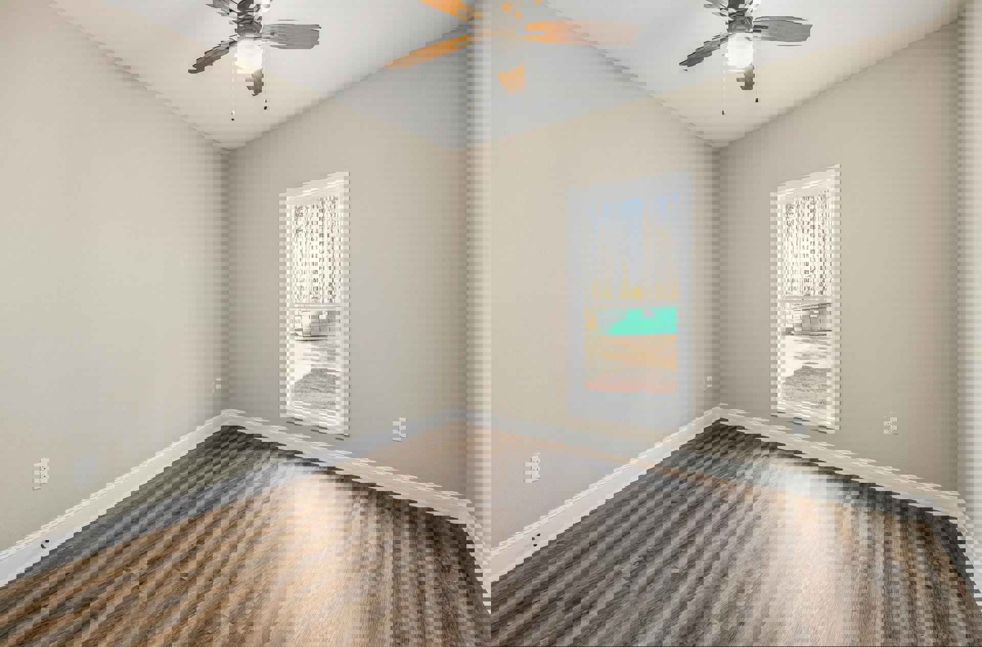 Sunlit room with wood laminate flooring, white plaster walls, ceiling fan with light, large window overlooking a green storage bin outside