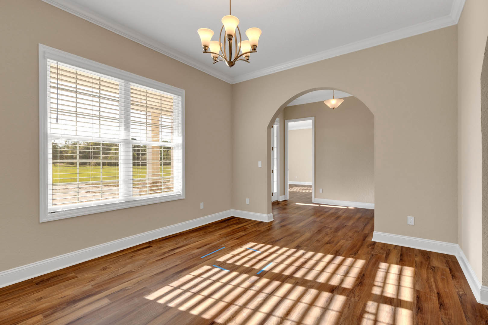 Wood floor with blue accent lines, white walls with decorative molding, window with white blinds, ceiling-mounted chandelier with multiple lights, sunlight casting shadows across