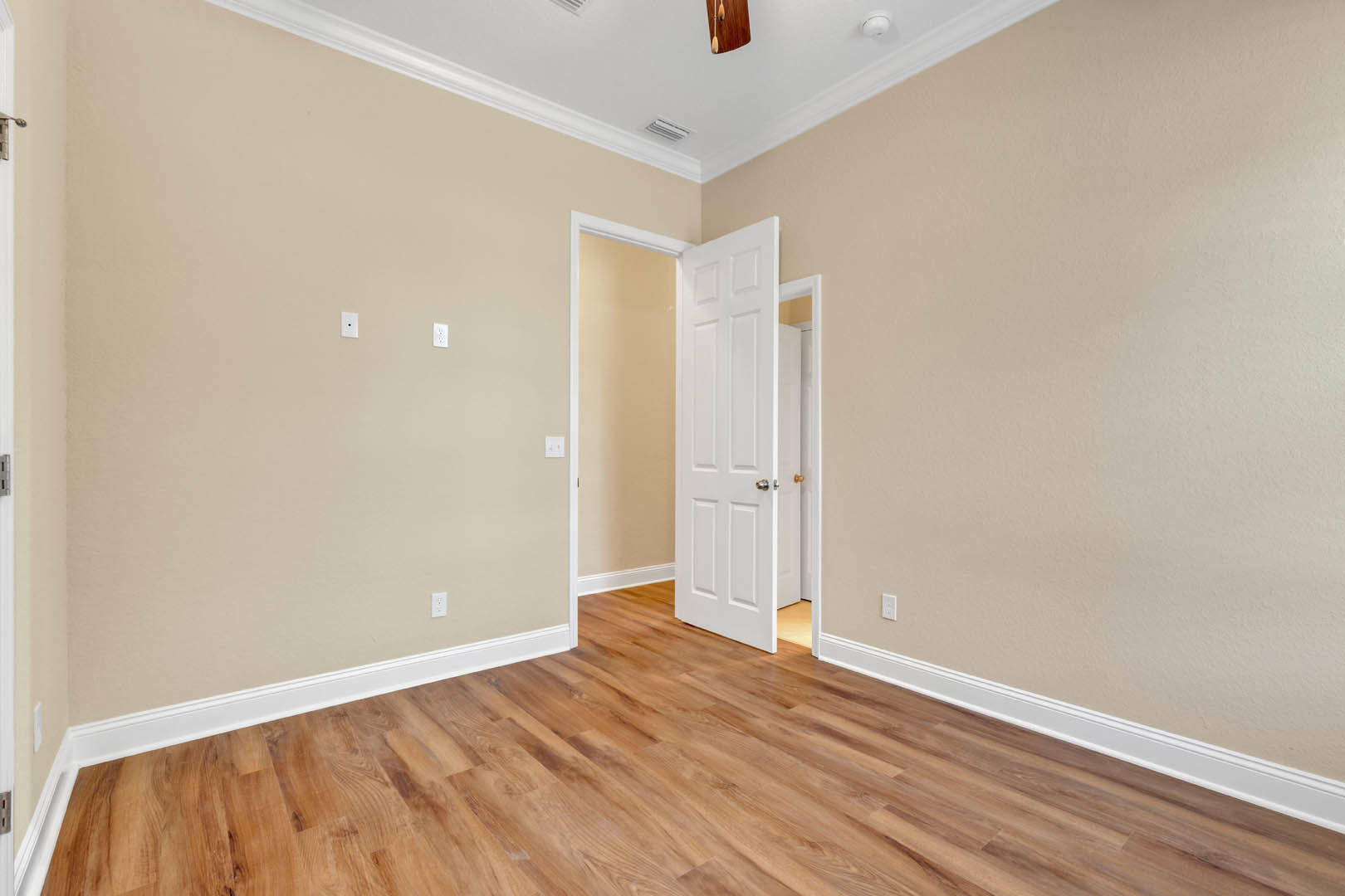 Wood floor room with white walls, open white door with silver handle, ceiling fan, wall-mounted switches, and a clock visible.