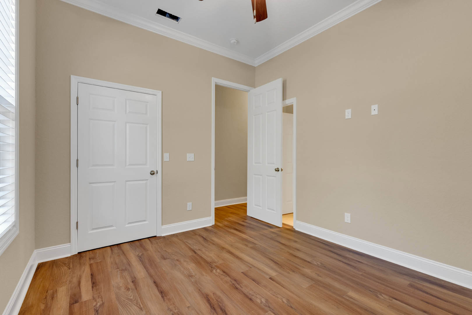 Room with hardwood flooring, white paneled doors featuring silver and gold handles, neutral walls, and ceiling fan.