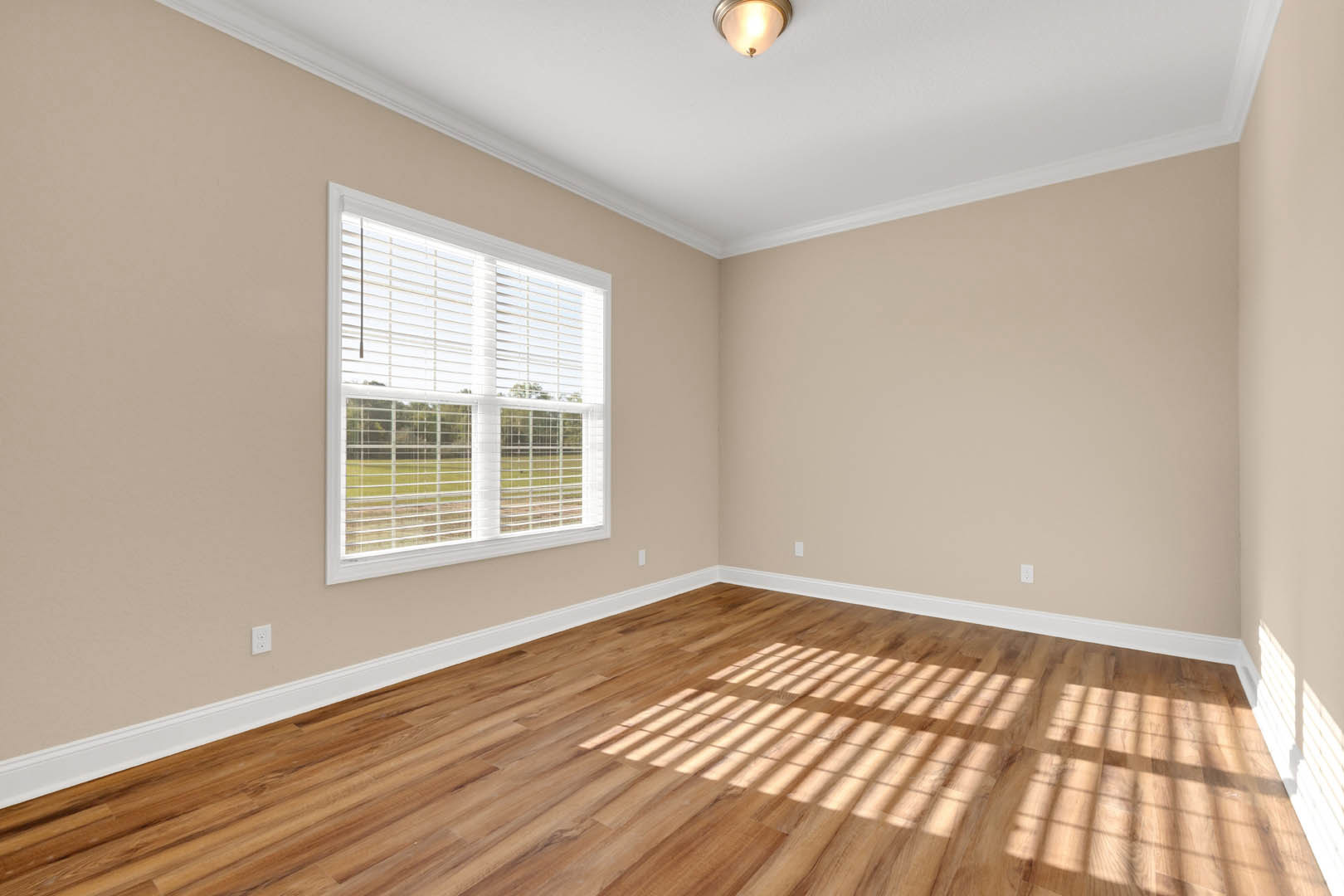 Sunlit room featuring hardwood flooring, white walls, large window with closed blinds, ceiling light fixture, and crown molding.