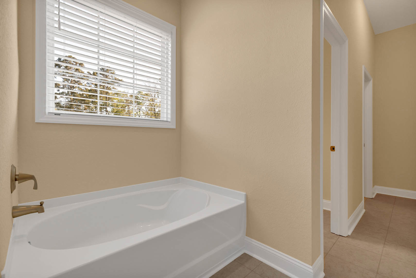 White freestanding bathtub beneath a large window with horizontal blinds, chrome faucet, light gray tile flooring, and neutral painted walls