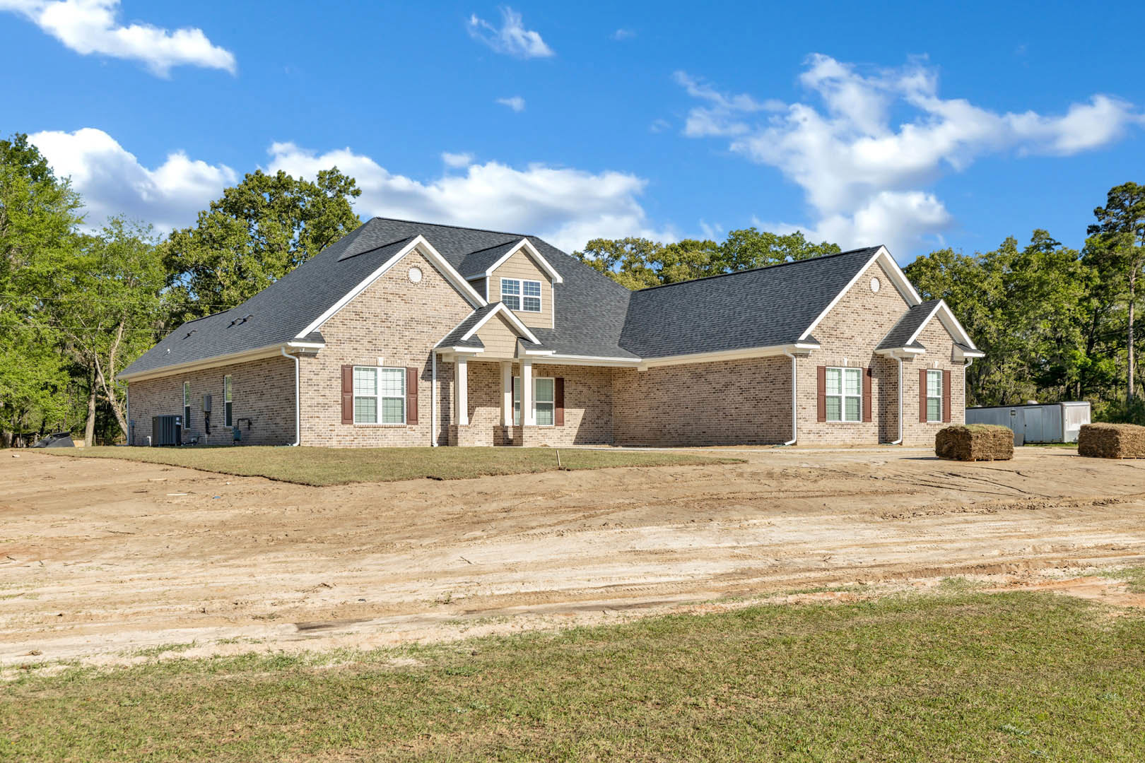 Brick house with grey roof, white-trimmed windows, and a dirt road bordered by grass under a blue sky with scattered clouds; bale of hay visible in foreground.