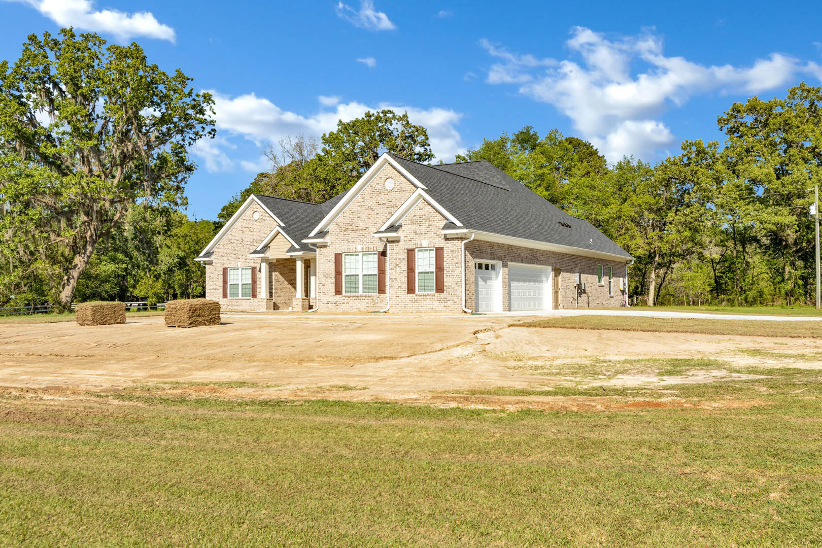 Two-story farmhouse with white roof, attached garage, expansive green lawn, mature tree with hanging moss, dirt driveway, and large hay bale in foreground