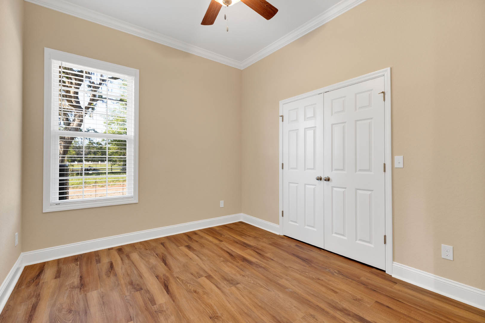 Wood flooring with white baseboards, white double doors, window with closed blinds, white electrical outlet on wall, partial view of ceiling.
