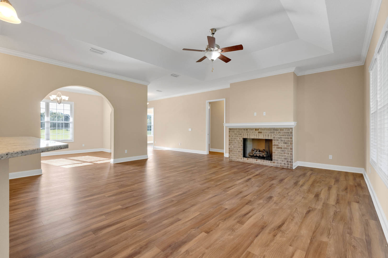 Living room with hardwood flooring, brick fireplace with stacked logs, ceiling fan with light fixture, window with white blinds, and white door frame
