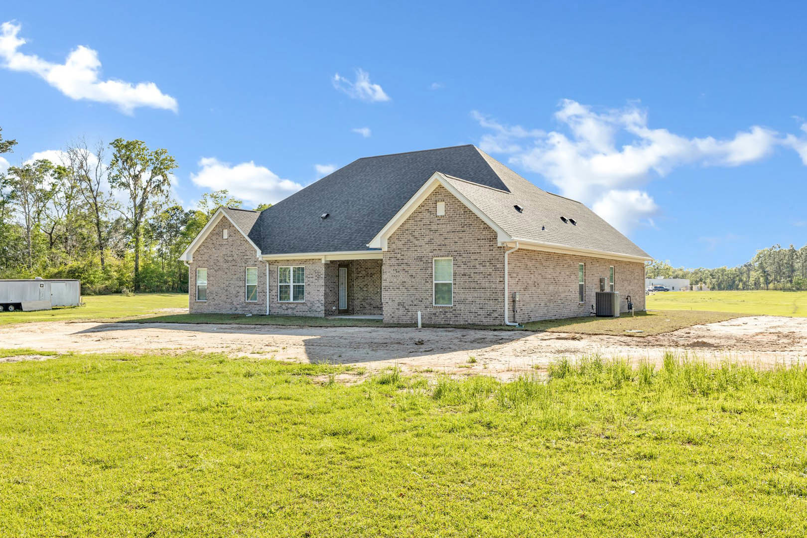 Red brick house with white roof and white-framed windows, set on a grassy lot with a dirt driveway, surrounded by trees and open sky.