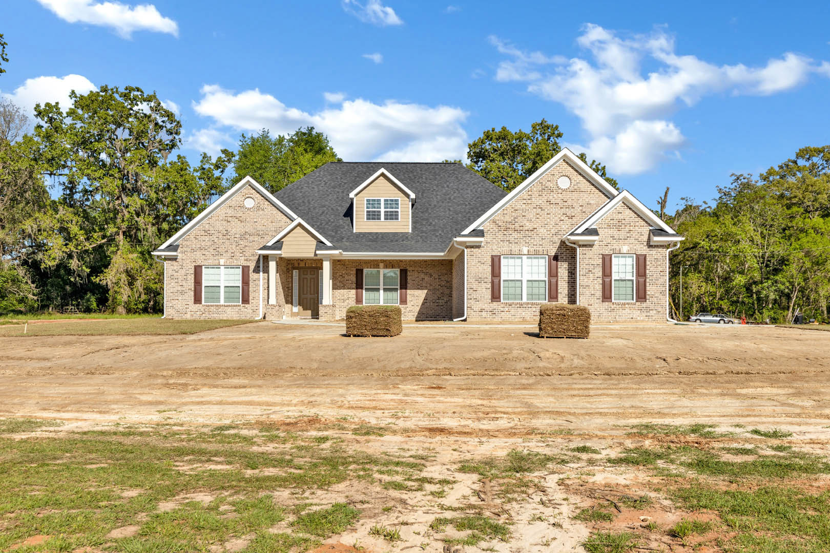 Modern house with black roof, large windows, and light siding, set beside a dirt road with grassy hill and trees in the background; pile of hay visible in foreground.