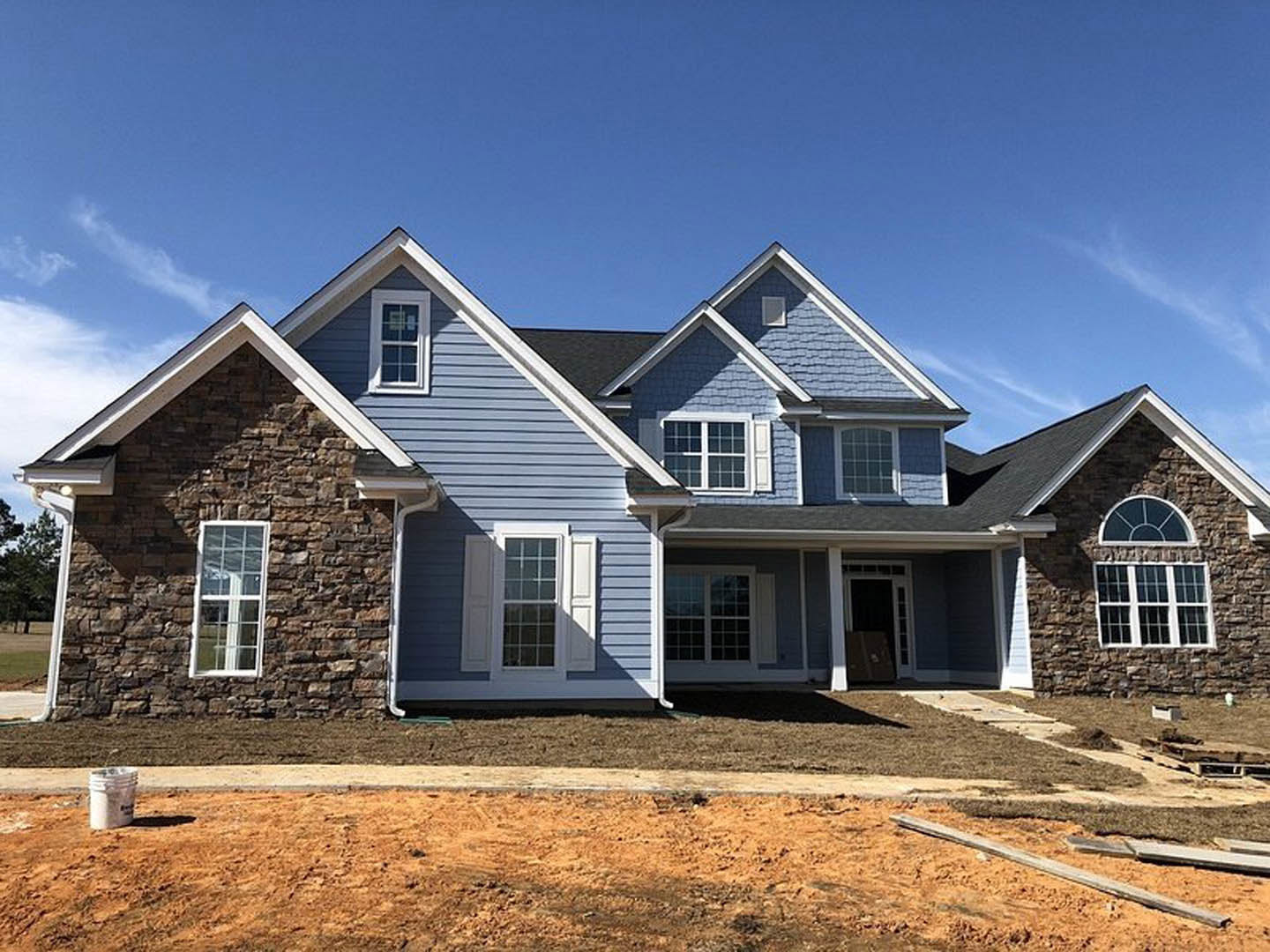 Partially built house with blue roof, white framed multi-pane windows, white buckets stacked near exterior, wooden plank lying on ground, clear blue sky overhead
