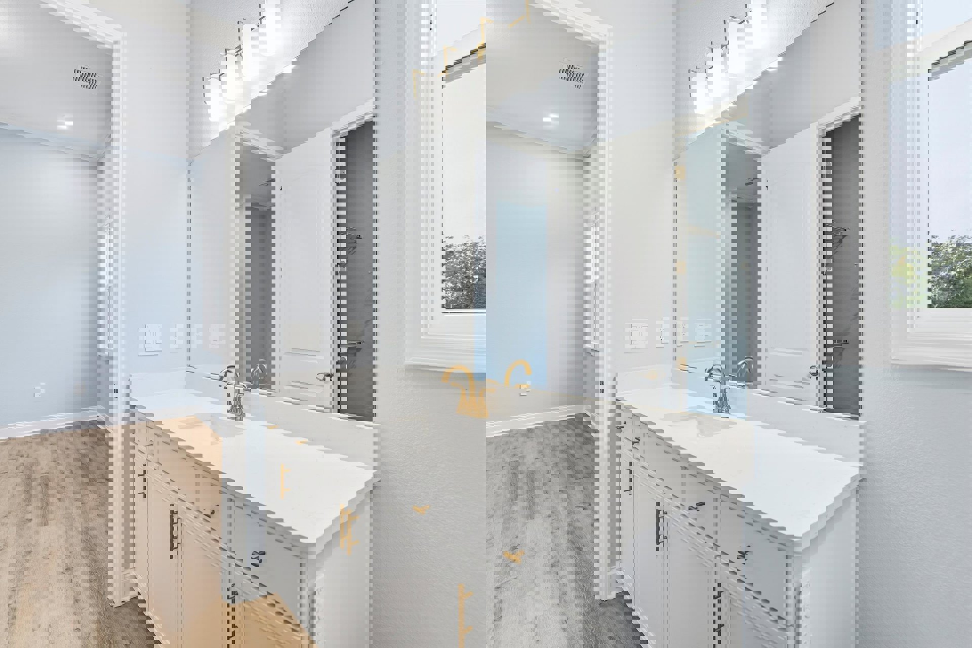 White bathroom with gold-handled sink, rectangular mirror above, metal wall shelf, wood flooring, white door with silver frame, trees visible through window in background