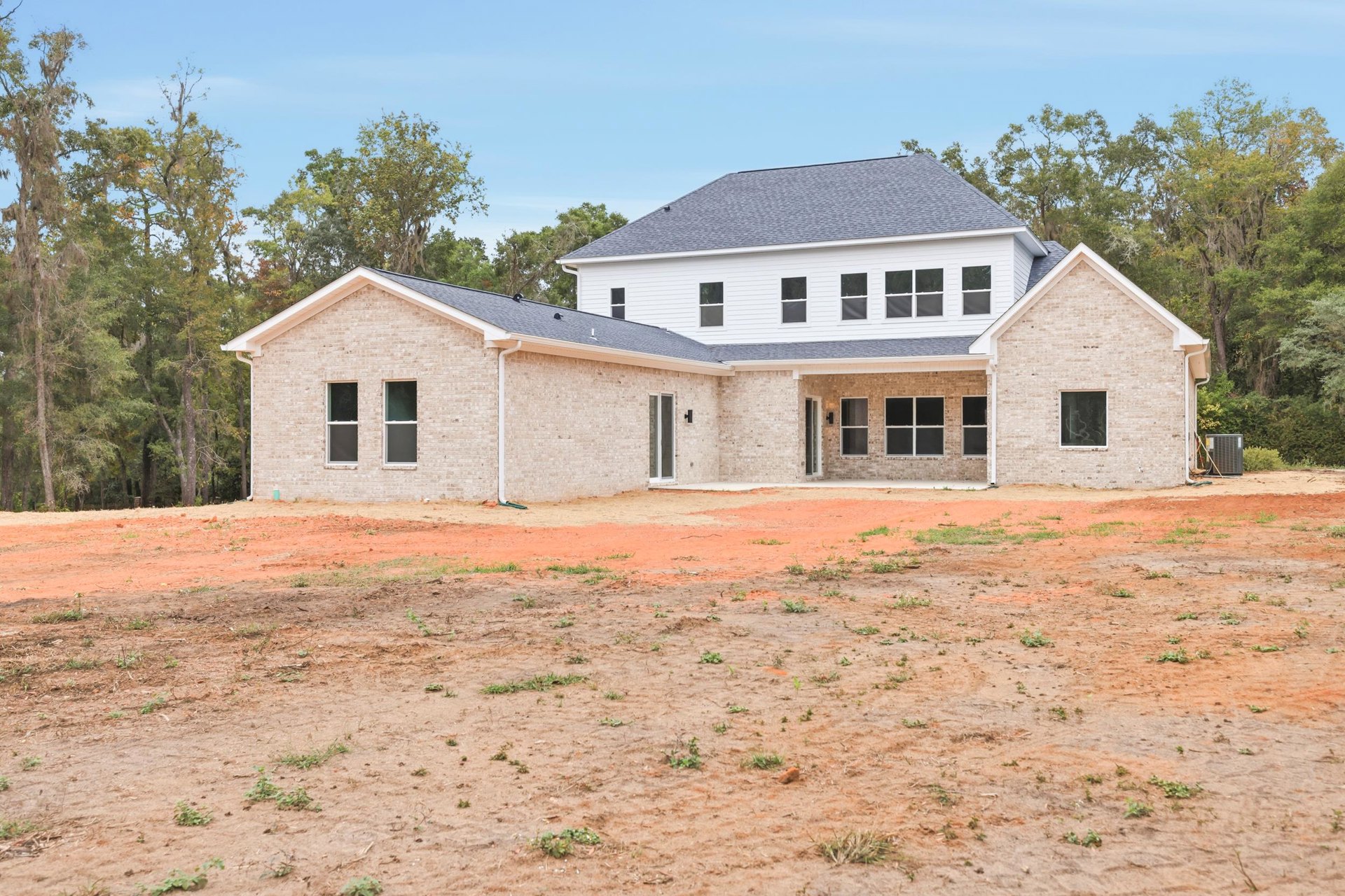 Single-story house with white roof and brick wall, cracked window, dirt yard in foreground, large metal object beside building, sparse trees and blue sky in background.