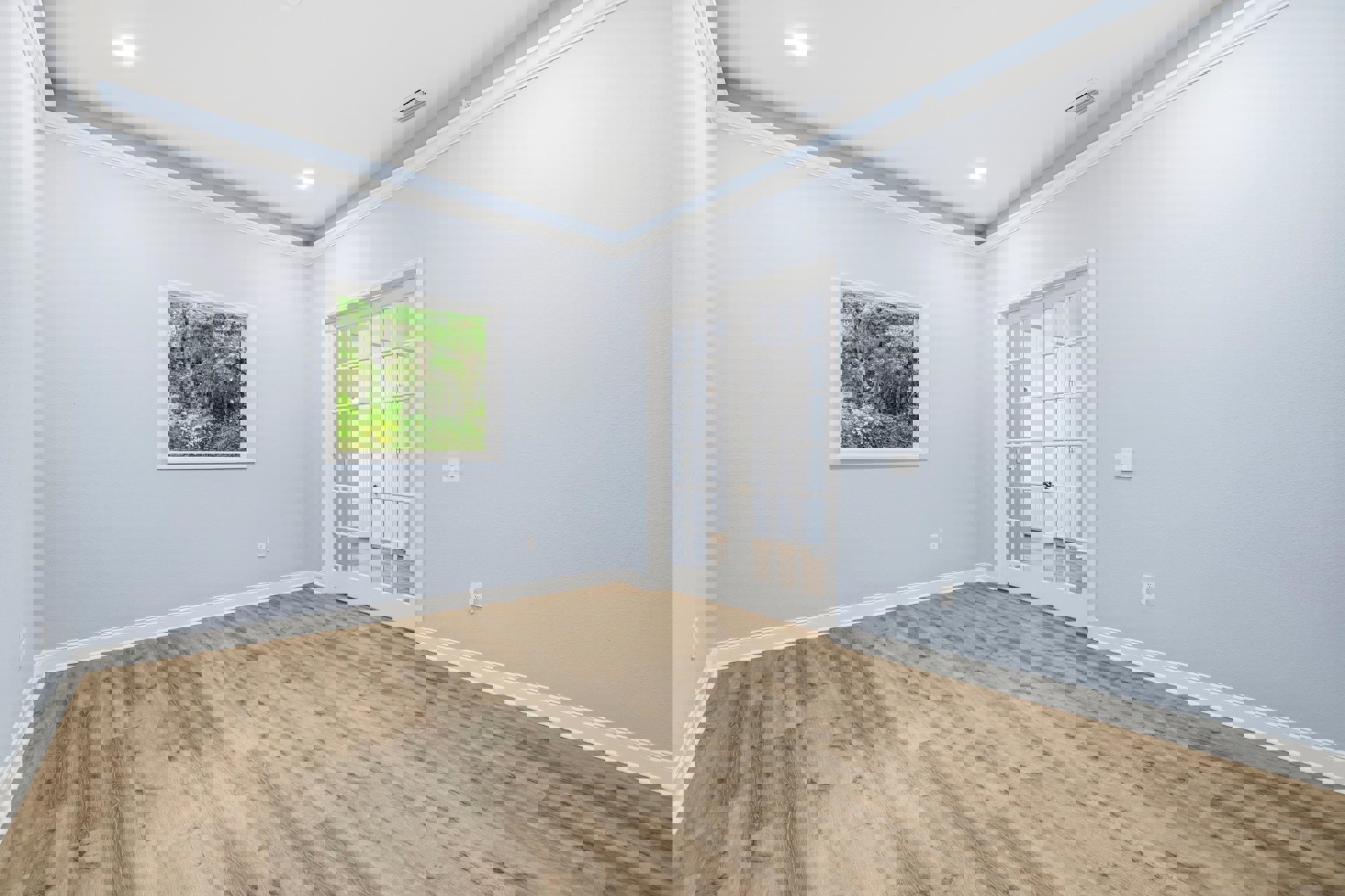 Wood floor room with white double doors featuring glass panels, large window overlooking trees, white vent with black stripes, and white ceiling with recessed light.